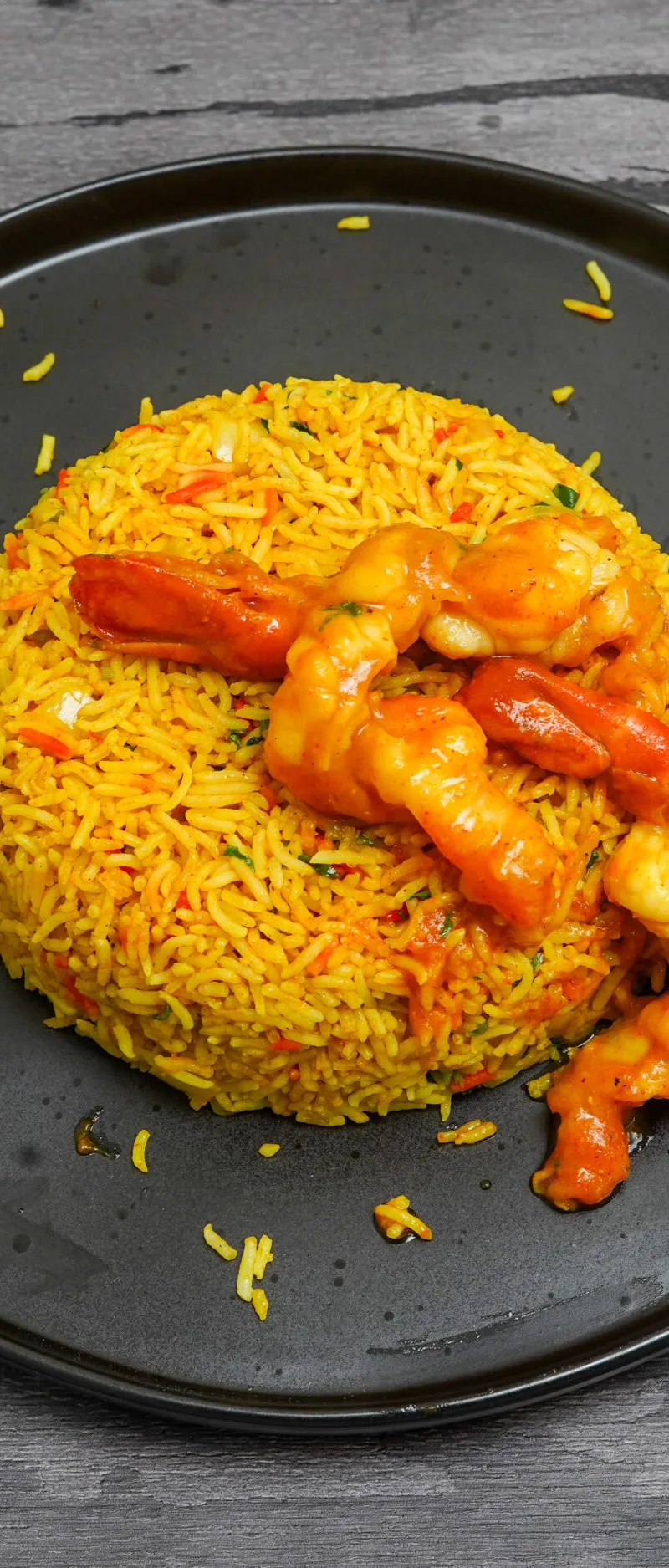 Bright yellow king prawn biryani in a black dish on a dark wooden surface, garnished with chili.