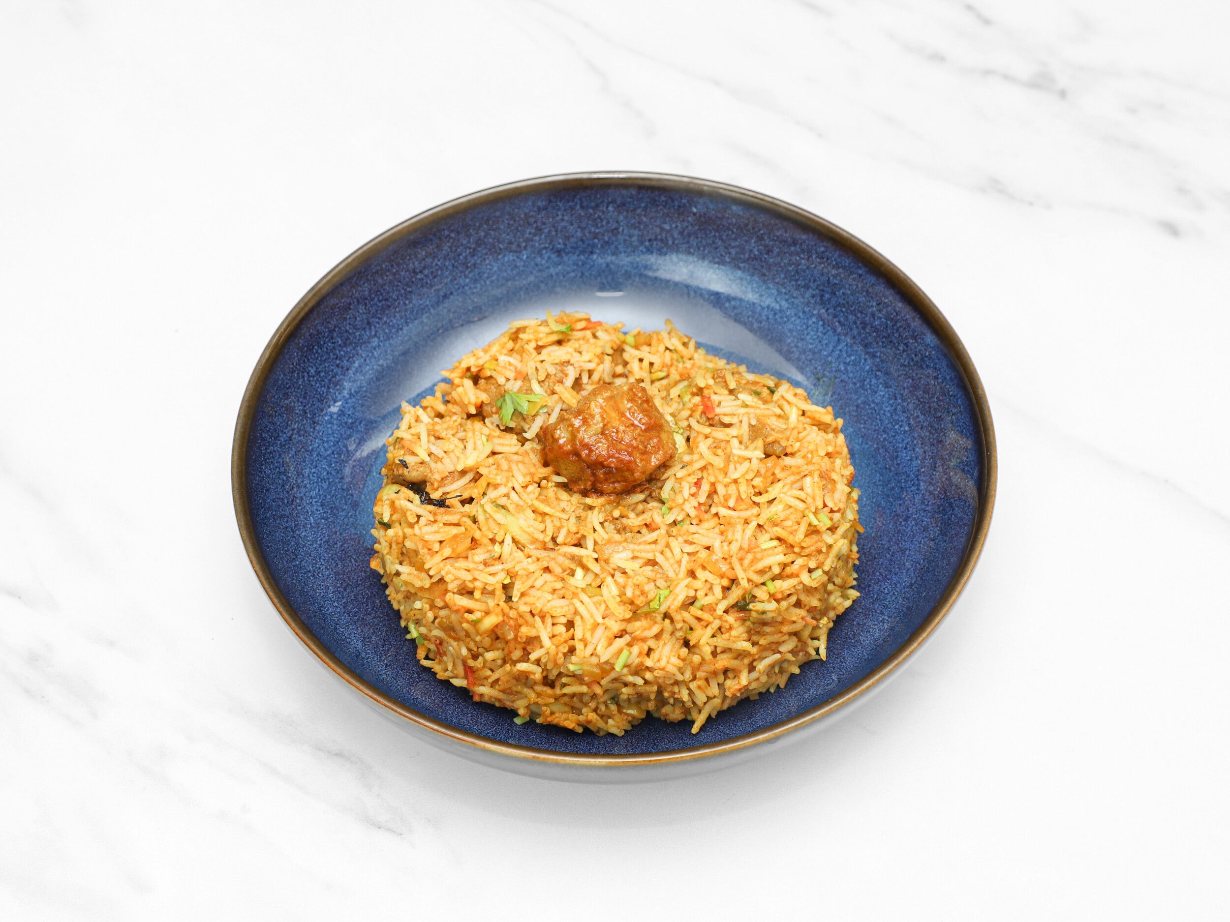 A blue bowl holds a generous portion of colourful biryani rice with a piece of meat visible on top.