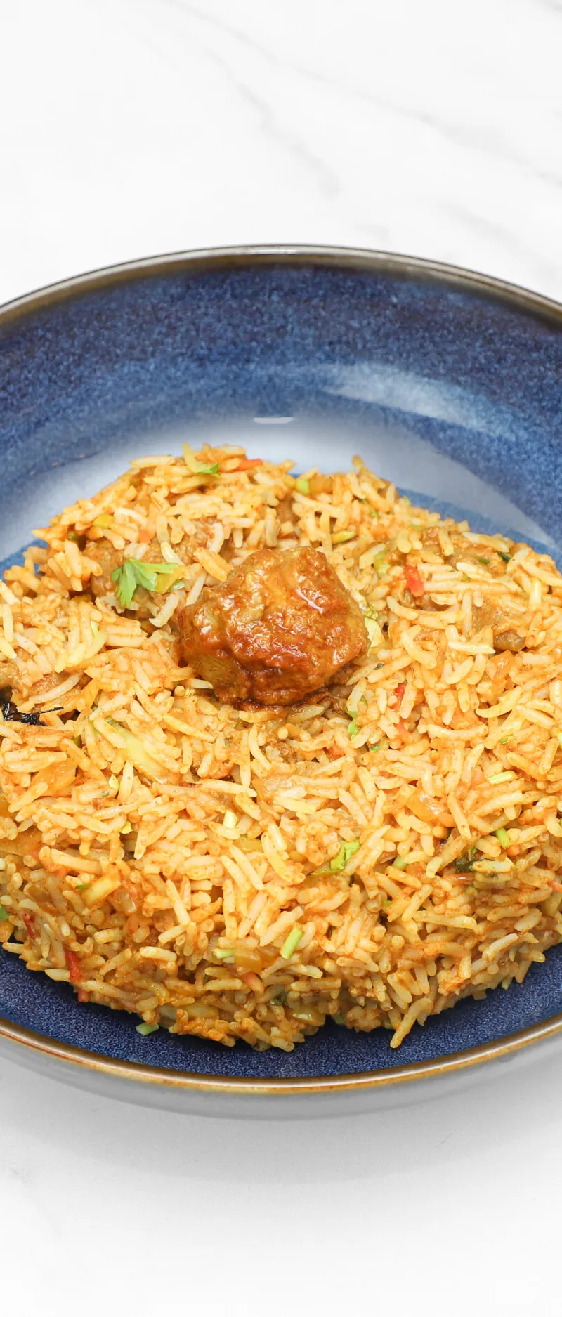 A blue bowl holds a generous portion of colourful biryani rice with a piece of meat visible on top.