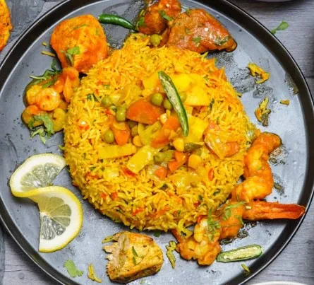 A black plate features vegetable biryani, accompanied by shrimps, paneer, and chicken, garnished with lemon and chili.