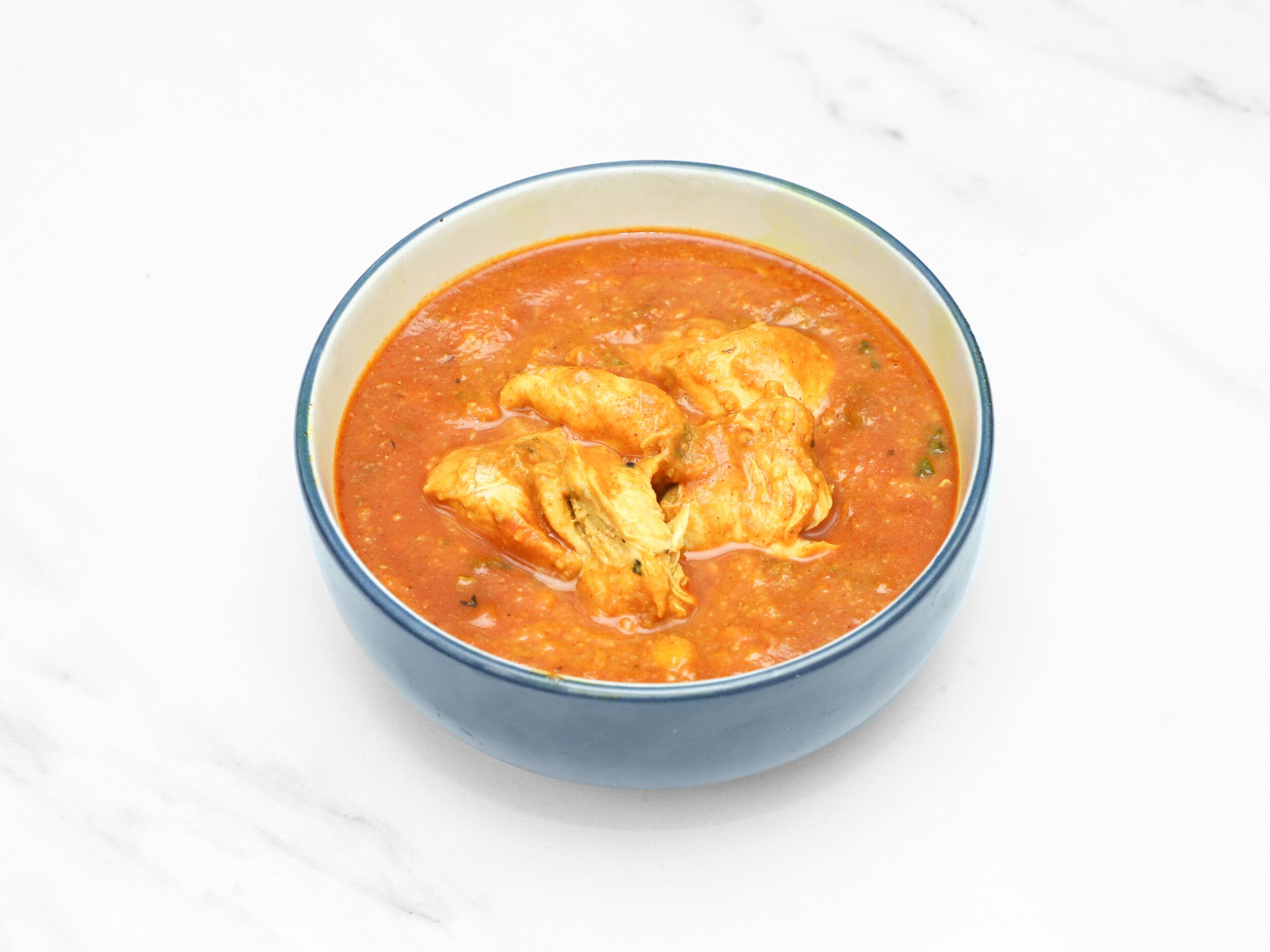 A blue bowl filled with chicken tikka masala on a white marble background.