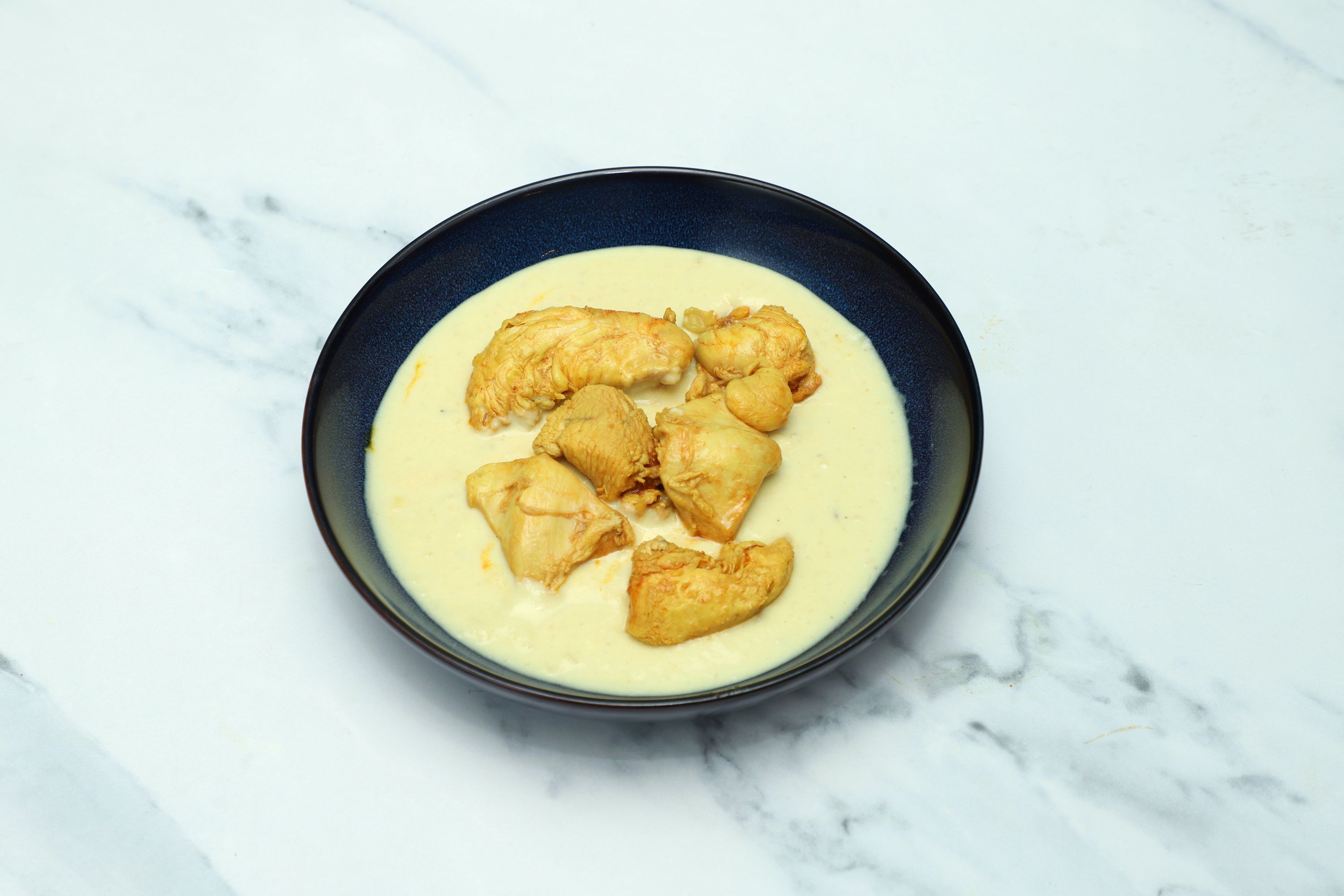 A dark blue bowl on a white marble surface filled with chicken pieces in a thick, light-yellow sauce.