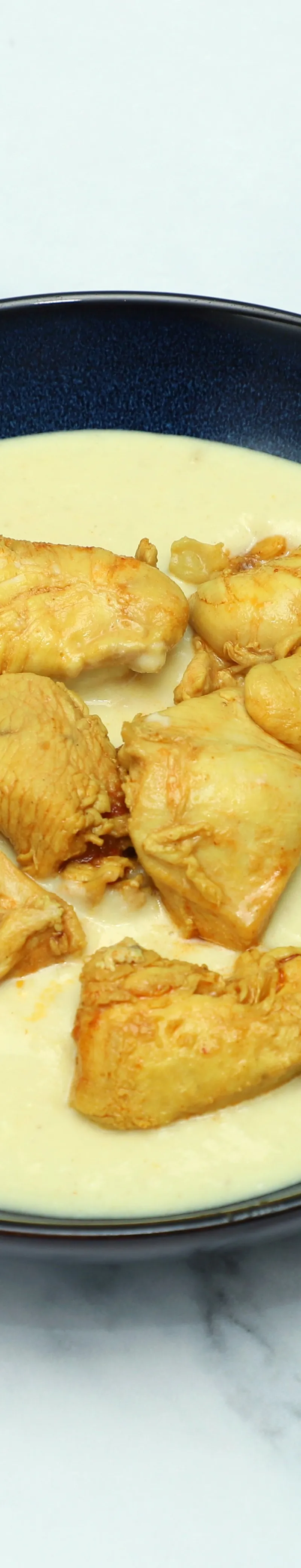 A dark blue bowl on a white marble surface filled with chicken pieces in a thick, light-yellow sauce.