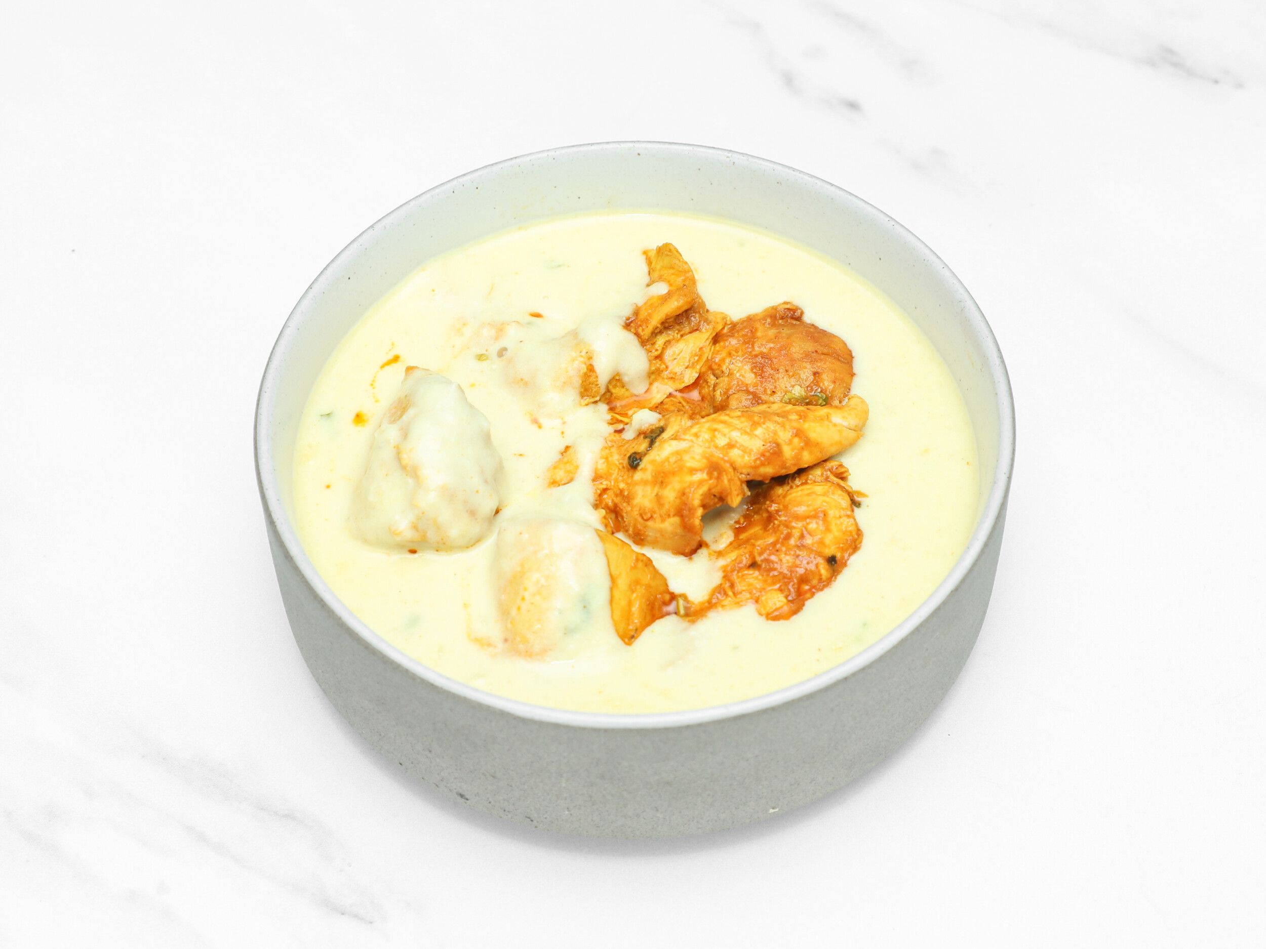 Creamy yellowish Malai chicken curry in a grey bowl on a white marble background