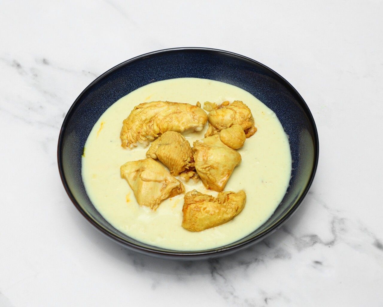 Chicken pieces in a pale yellow, creamy sauce served in a dark blue bowl on a white marble surface.