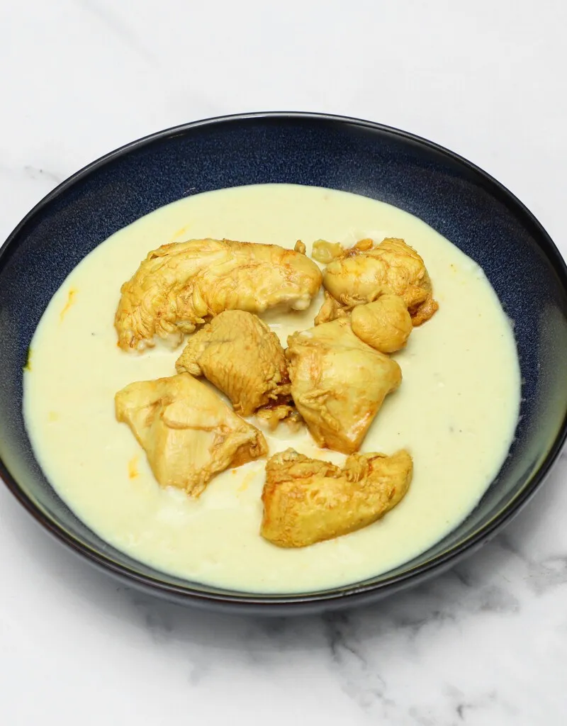 Chicken pieces in a pale yellow, creamy sauce served in a dark blue bowl on a white marble surface.