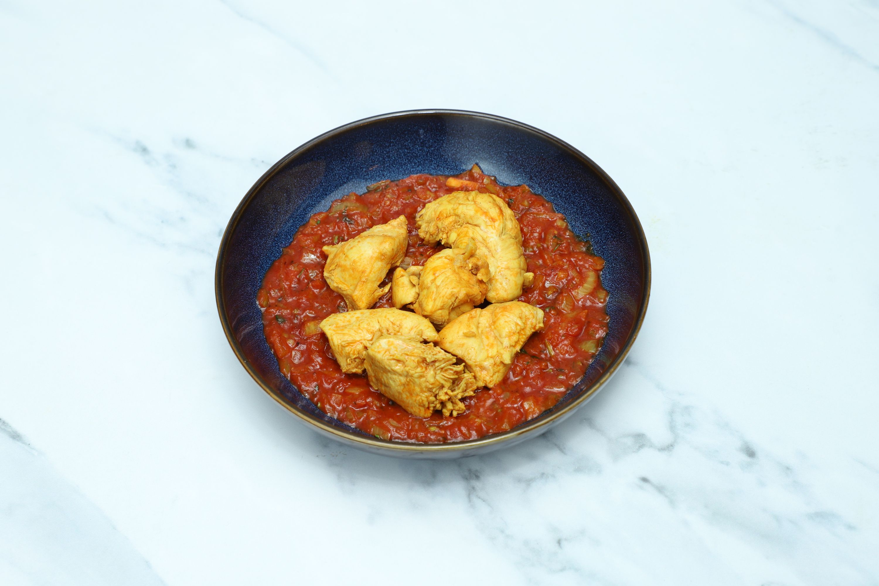 Golden chicken pieces nestled in a chunky red tomato and onion sauce in a dark blue bowl.