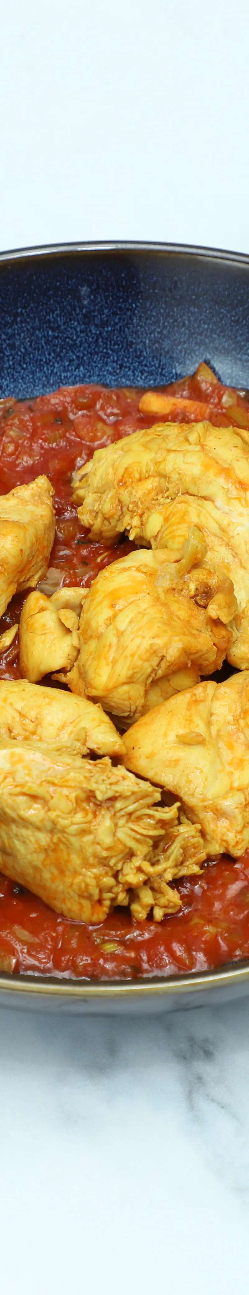 Golden chicken pieces nestled in a chunky red tomato and onion sauce in a dark blue bowl.