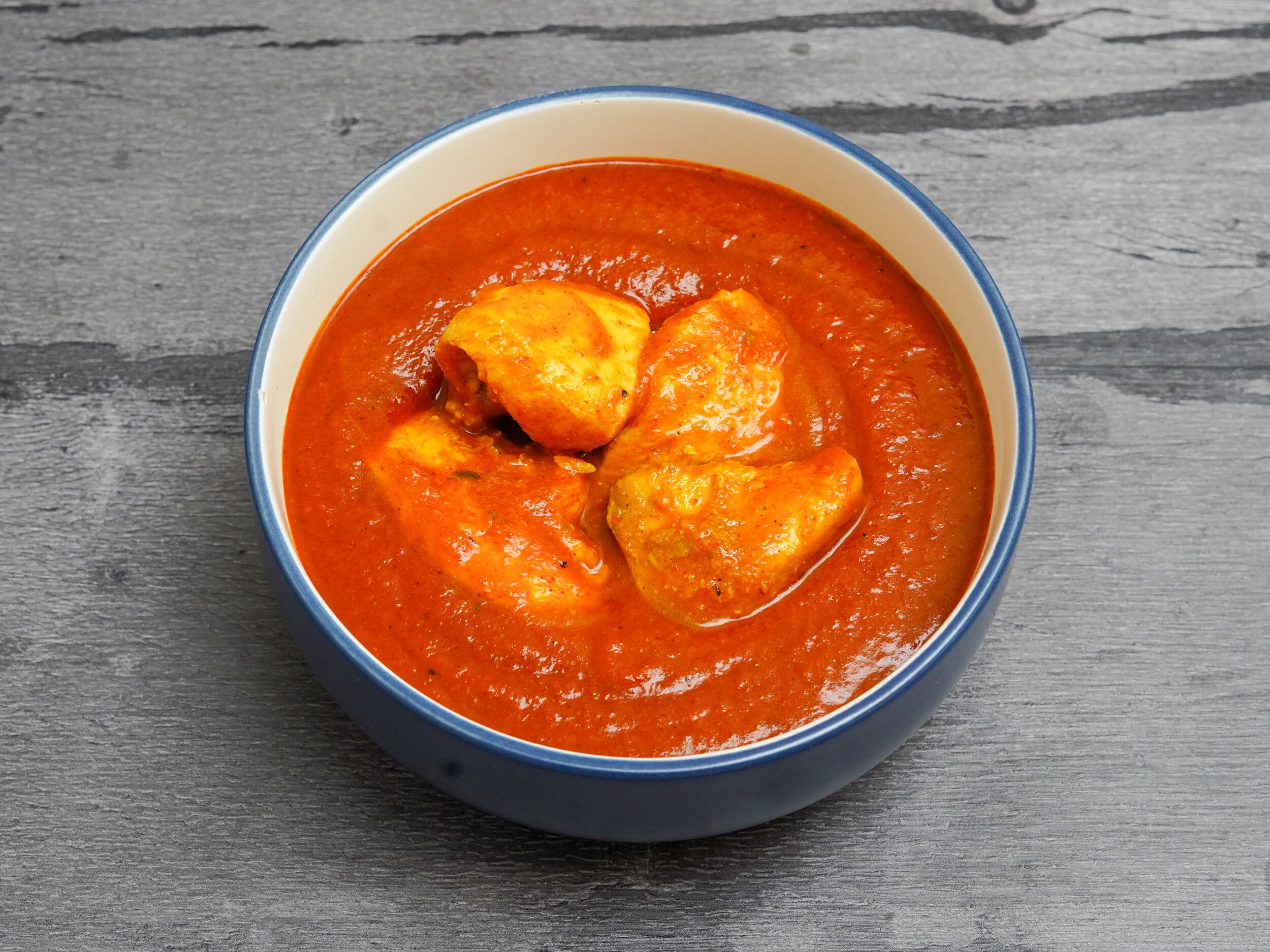 Chicken curry in a blue and white bowl, with chunks of chicken in a rich red sauce, against a grey wooden background.