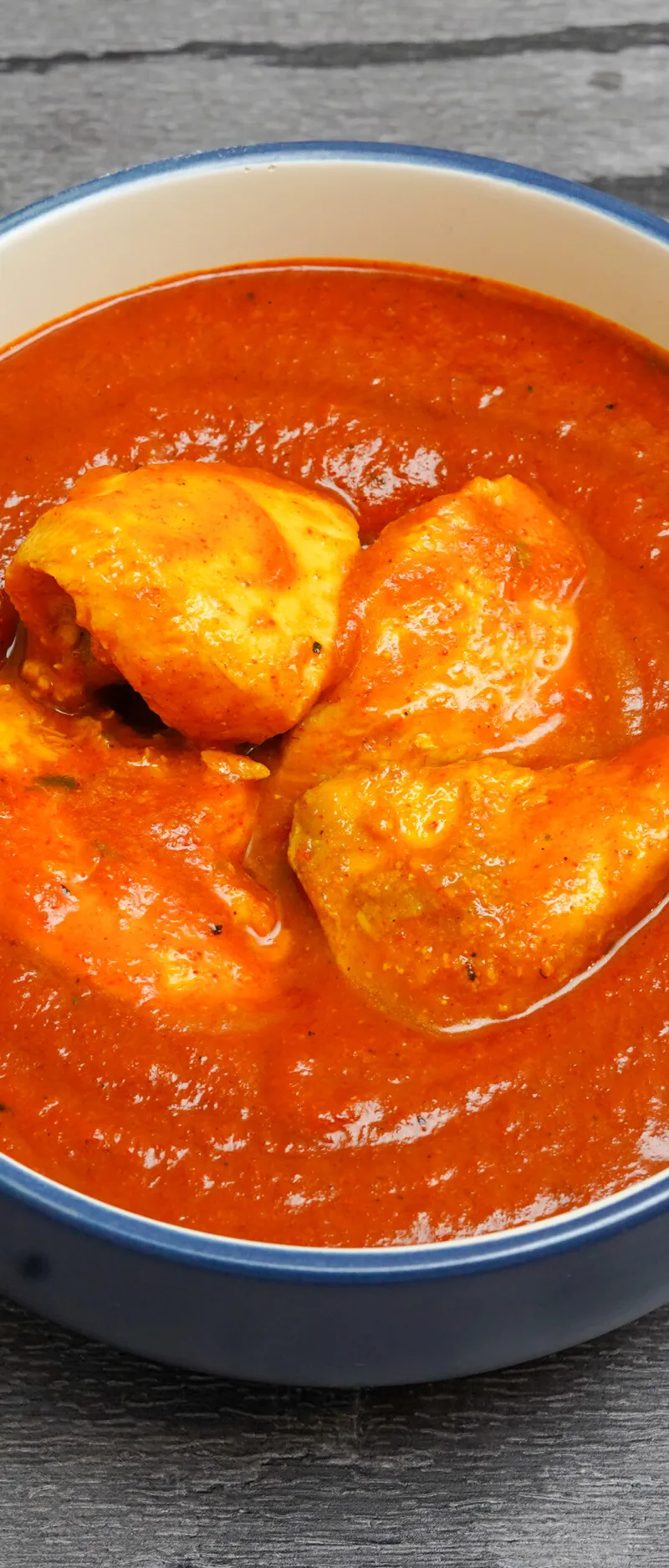 Chicken curry in a blue and white bowl, with chunks of chicken in a rich red sauce, against a grey wooden background.