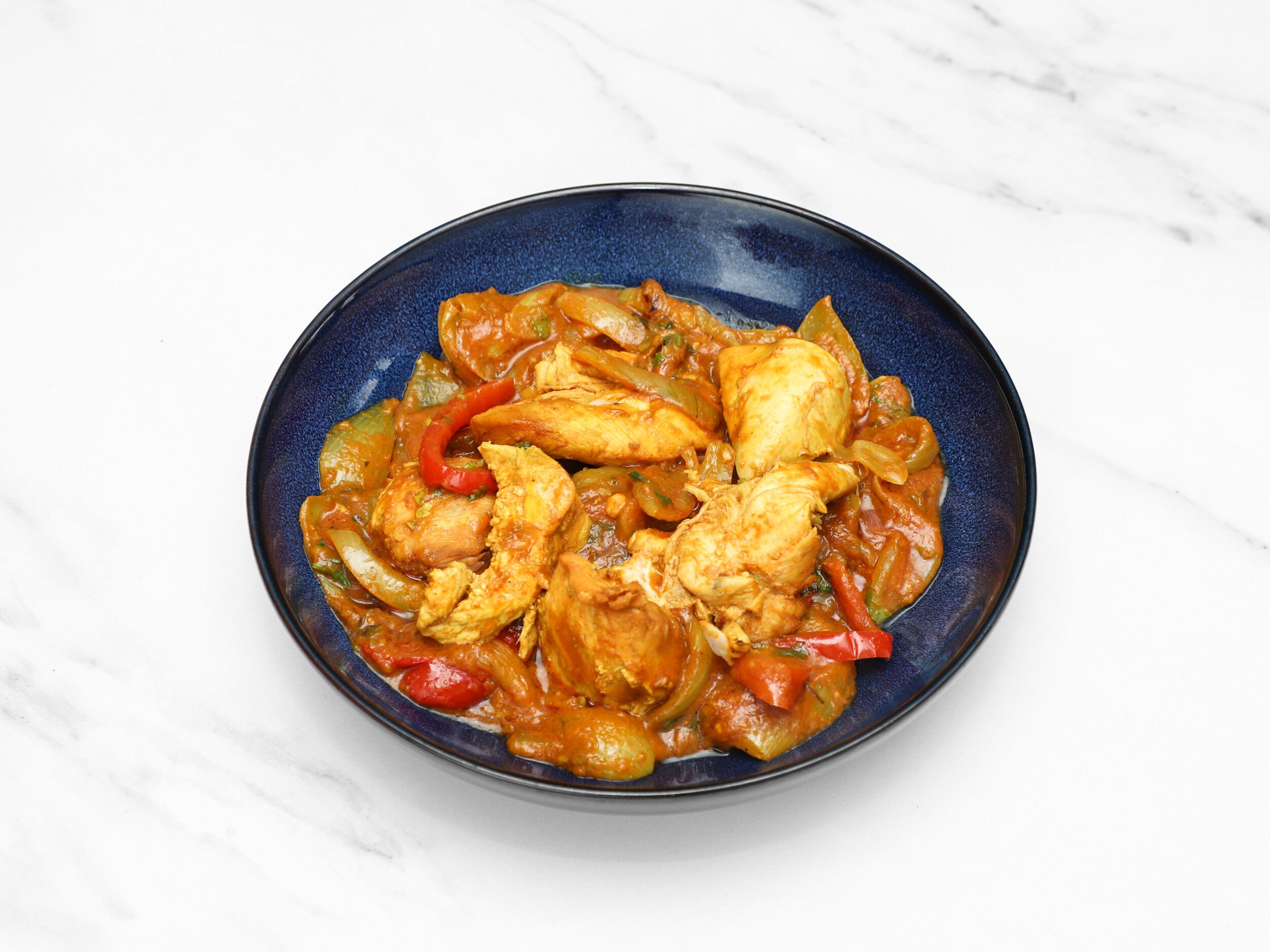 Chicken curry with large chunks of chicken, yellow onions, and red and green peppers in a rich sauce, served in a blue bowl.