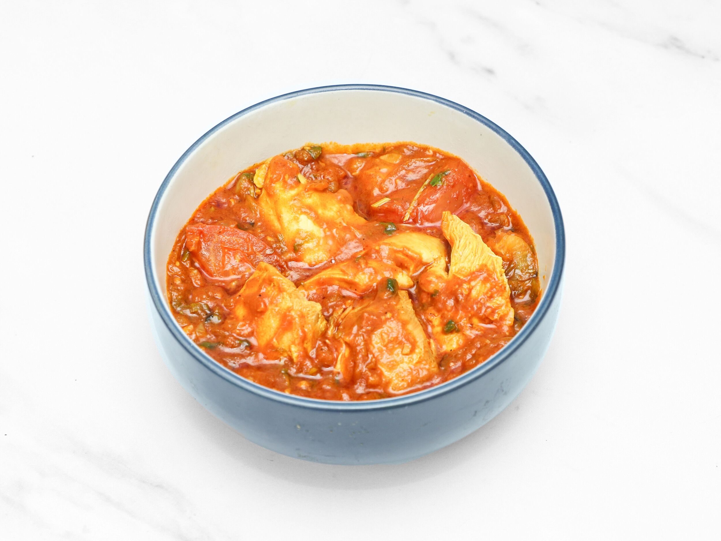 Chicken and tomato curry in a light blue ceramic bowl on a white marble surface.
