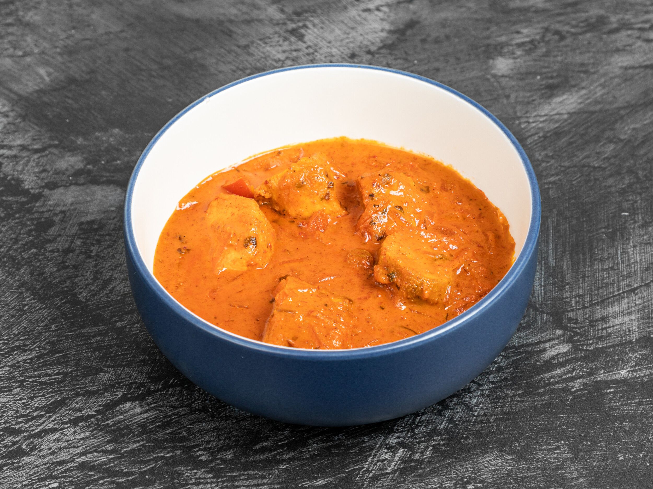 A blue and white bowl containing a vibrant orange chicken tikka masala curry with visible chicken pieces and tomato.