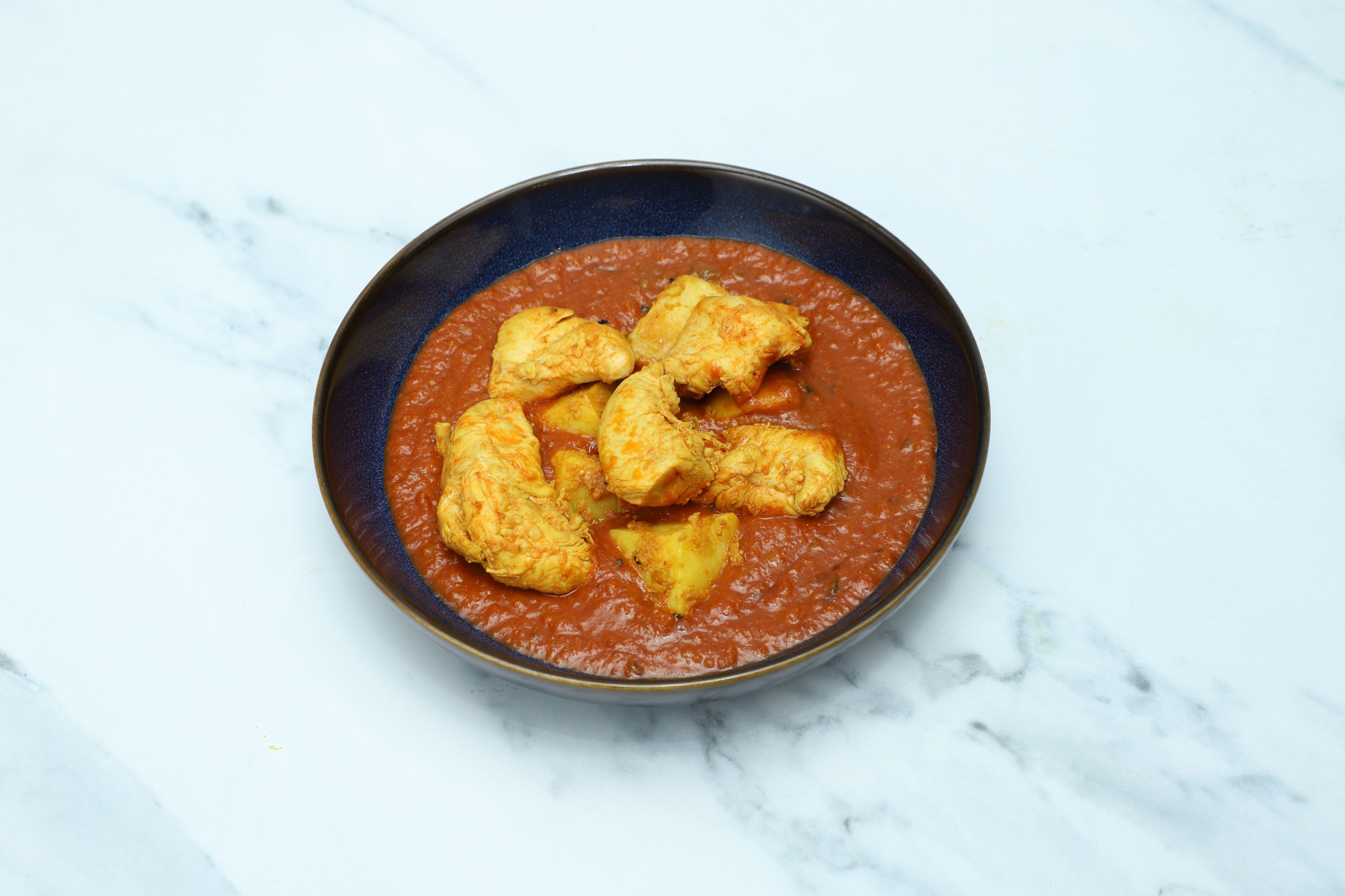 Aloo chicken curry with golden chicken pieces and potato chunks in a rich red sauce, served in a contemporary blue bowl on a marble surface.