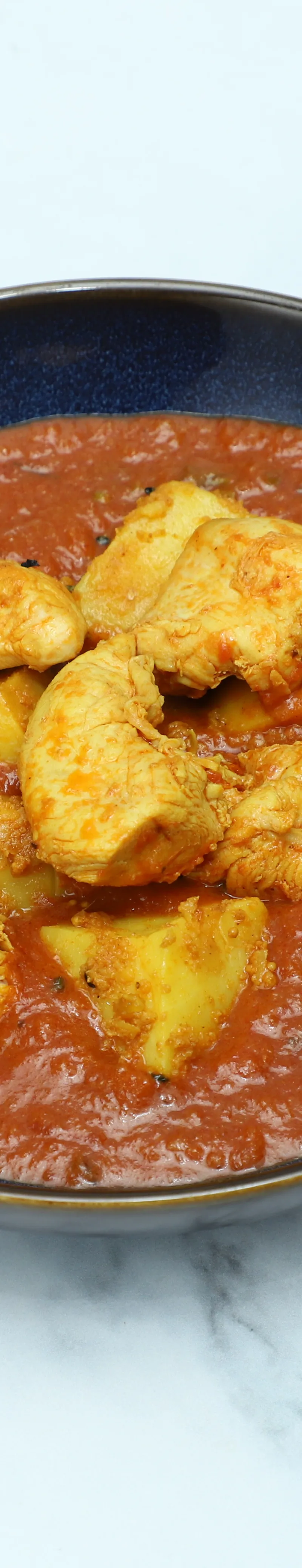 Aloo chicken curry with golden chicken pieces and potato chunks in a rich red sauce, served in a contemporary blue bowl on a marble surface.