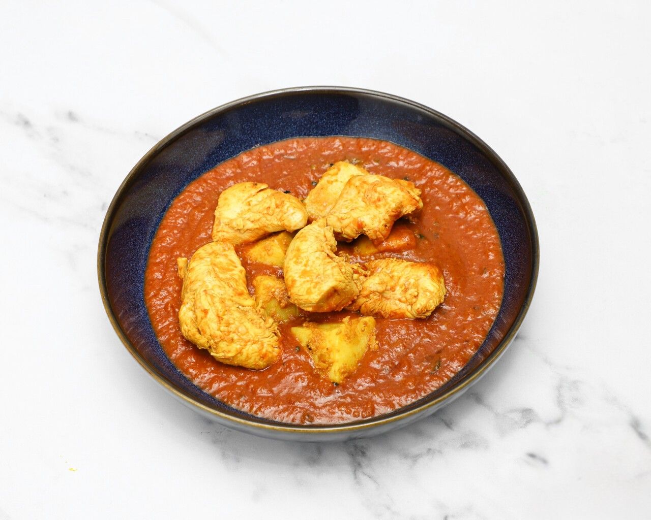 Chicken and potato pieces in a richly coloured red-orange curry sauce, in a dark blue bowl.
