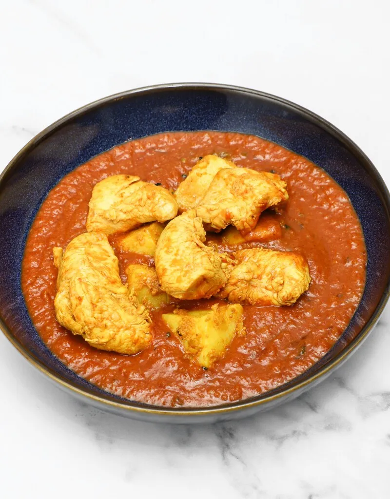 Chicken and potato pieces in a richly coloured red-orange curry sauce, in a dark blue bowl.