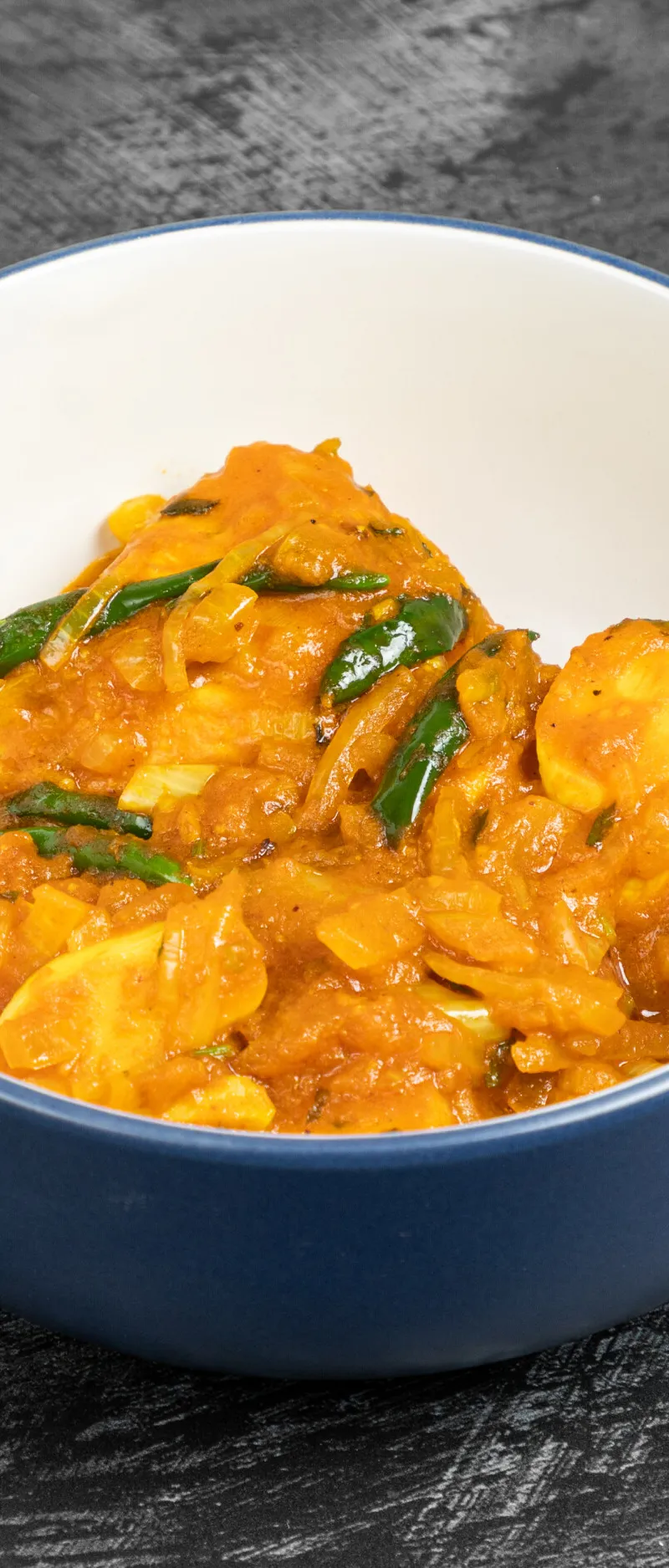 Aloo shim, an Indian potato dish in an orange sauce with sliced green chillies, served in a blue bowl with a white rim