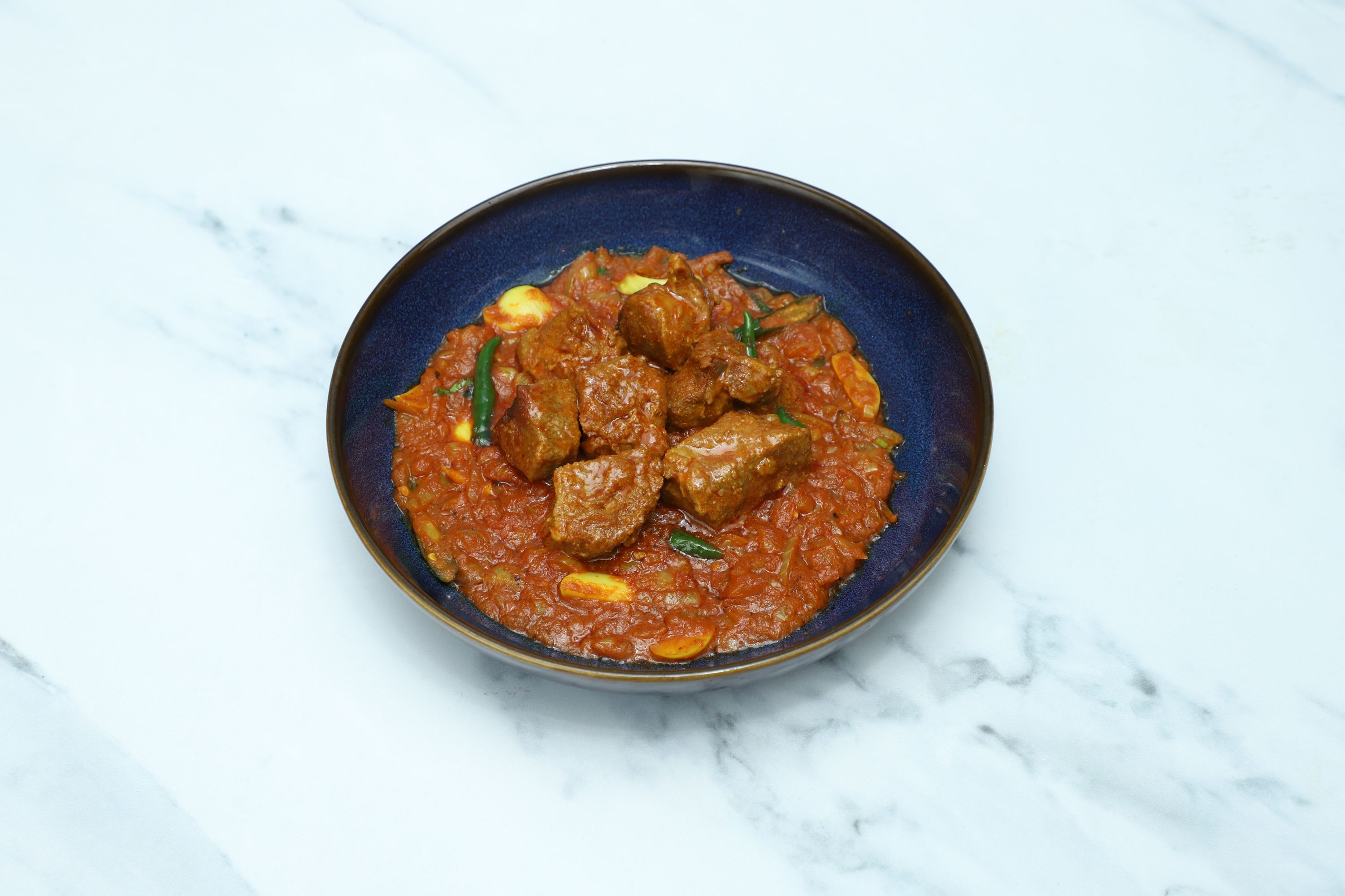 A round, blue bowl filled with lamb curry, featuring tender meat pieces and green chillies.