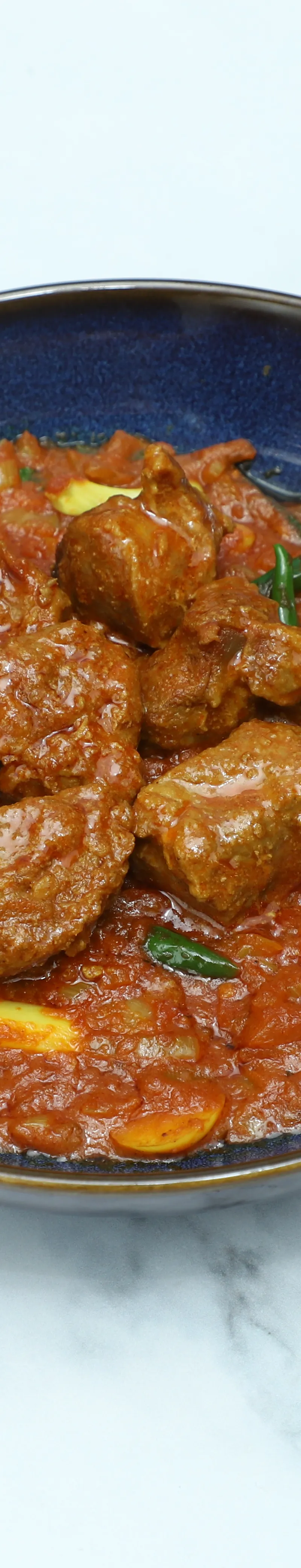 A round, blue bowl filled with lamb curry, featuring tender meat pieces and green chillies.