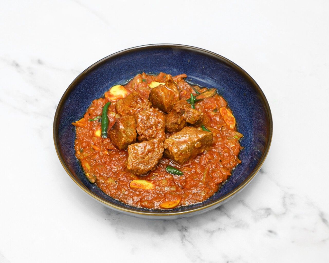Tender pieces of lamb curry in a rich red sauce, garnished with green chilies, served in a deep blue bowl