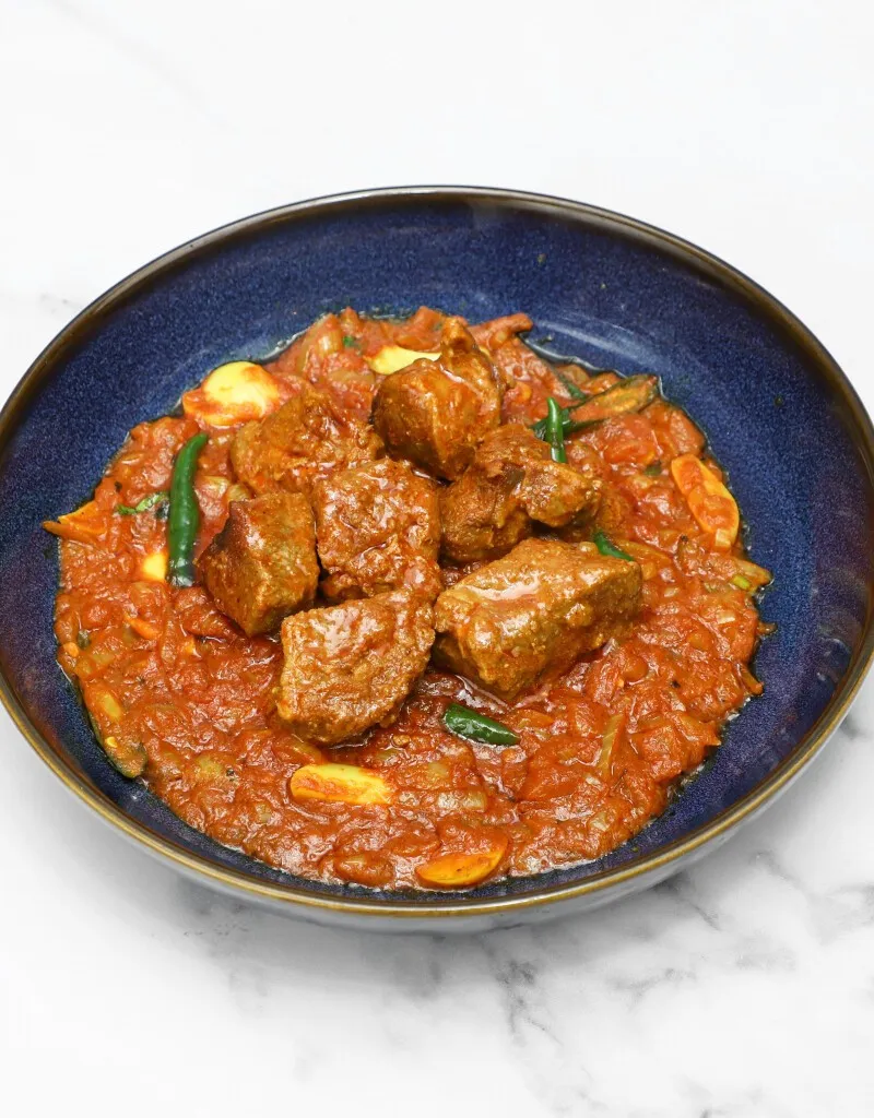 Tender pieces of lamb curry in a rich red sauce, garnished with green chilies, served in a deep blue bowl