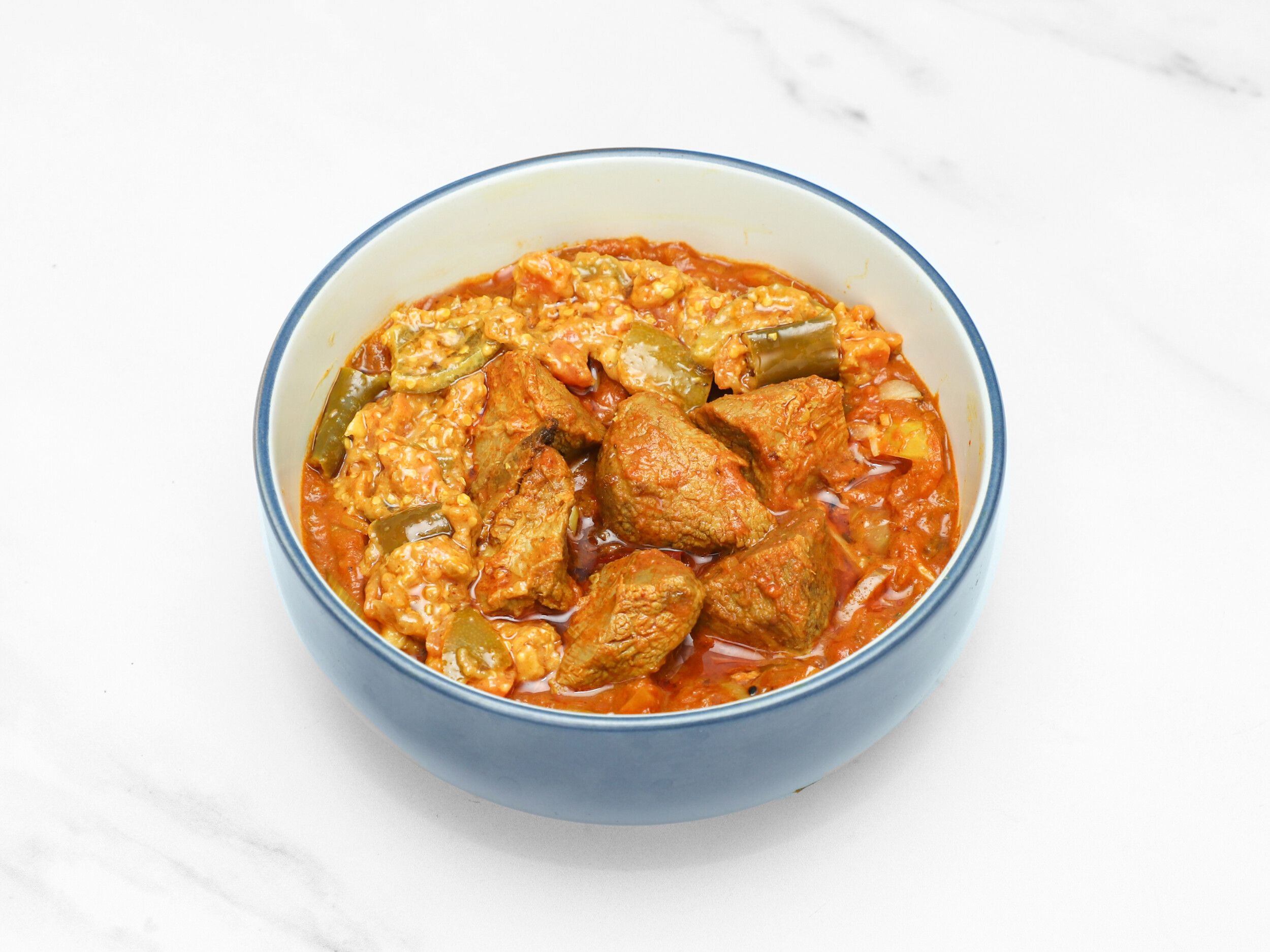 A close-up of a blue ceramic bowl filled with a rich red curry, chunks of beef, and green okra slices.