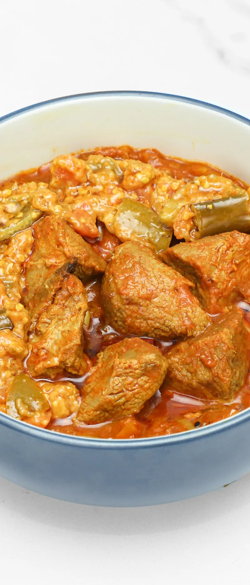 A close-up of a blue ceramic bowl filled with a rich red curry, chunks of beef, and green okra slices.