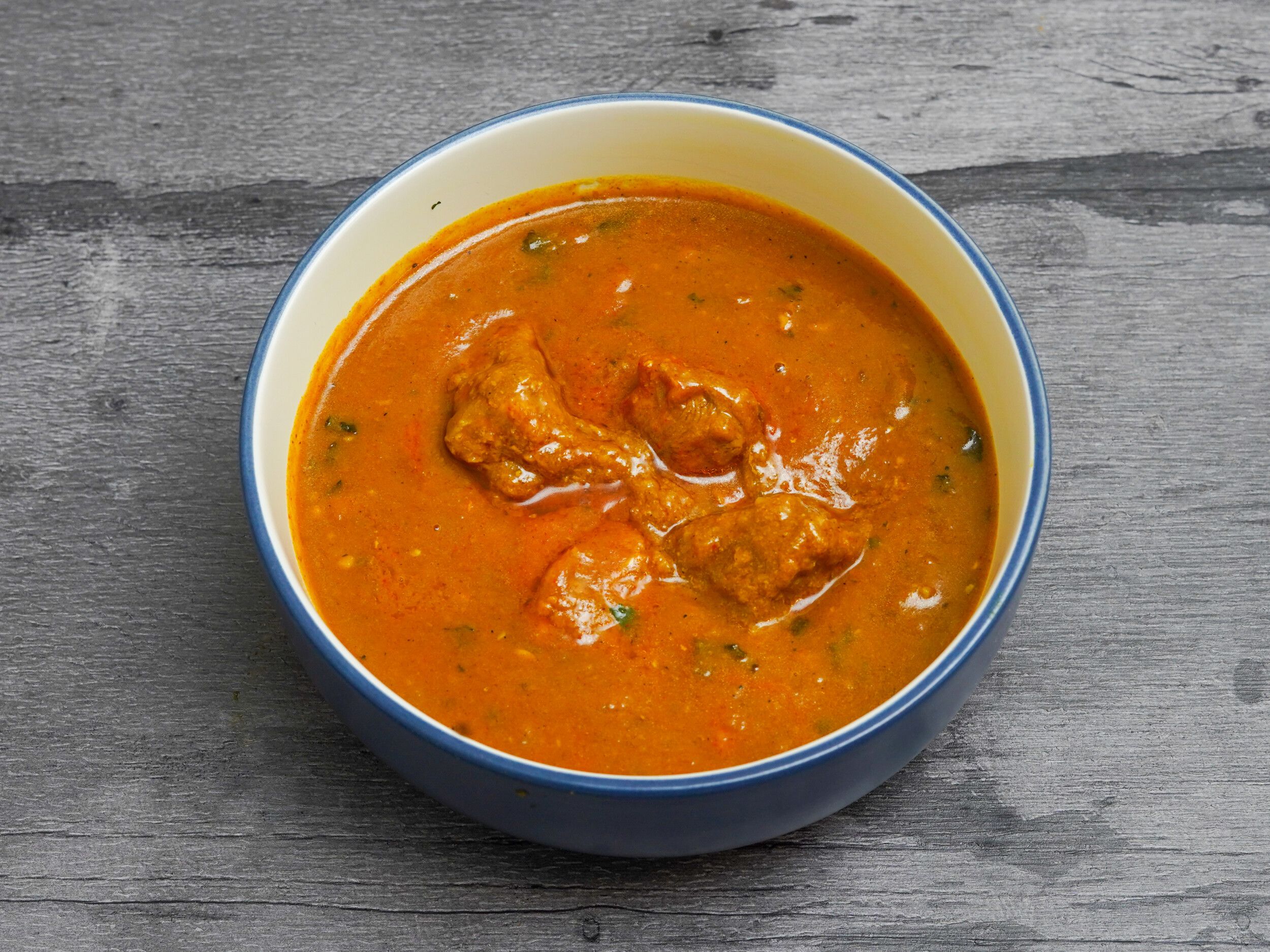 Top-down view of a blue and white bowl filled with orange chicken curry against a grey wooden background.
