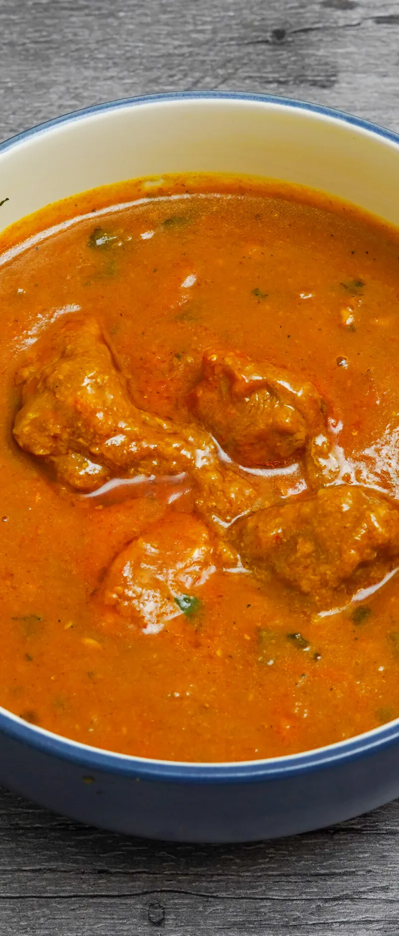 Top-down view of a blue and white bowl filled with orange chicken curry against a grey wooden background.