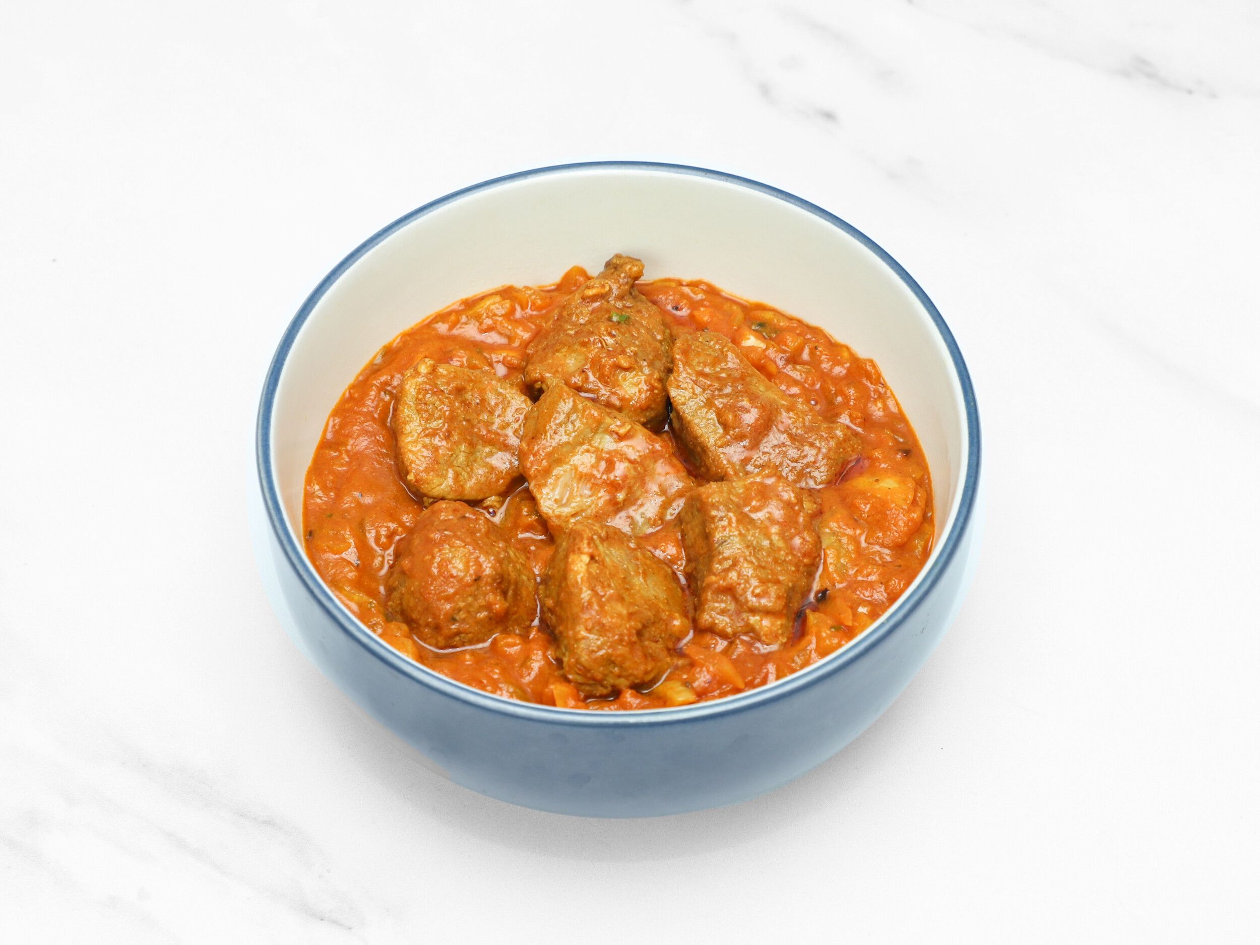 Meatballs in a rich orange-red curry sauce in a blue-rimmed white bowl on a white surface.