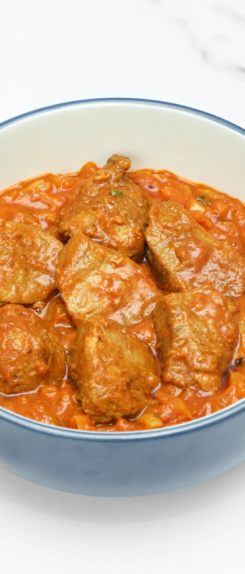 Meatballs in a rich orange-red curry sauce in a blue-rimmed white bowl on a white surface.