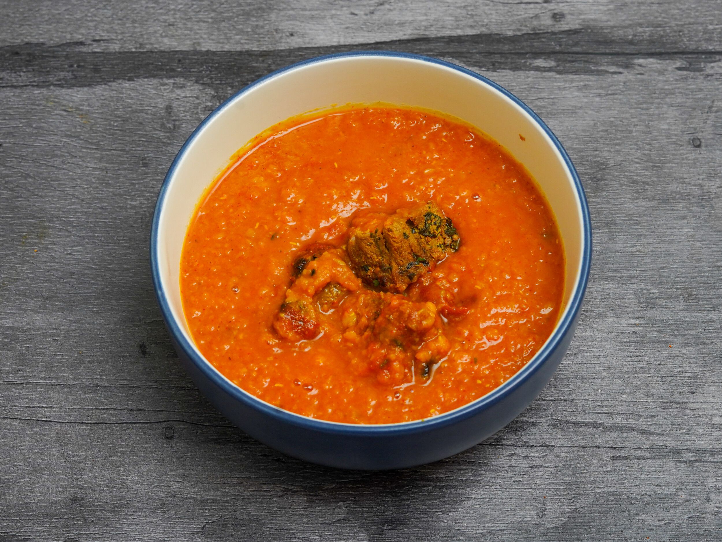 Bright orange lentil curry (dal) containing several kofta balls, served in a white bowl with a blue rim, on a dark wooden surface.