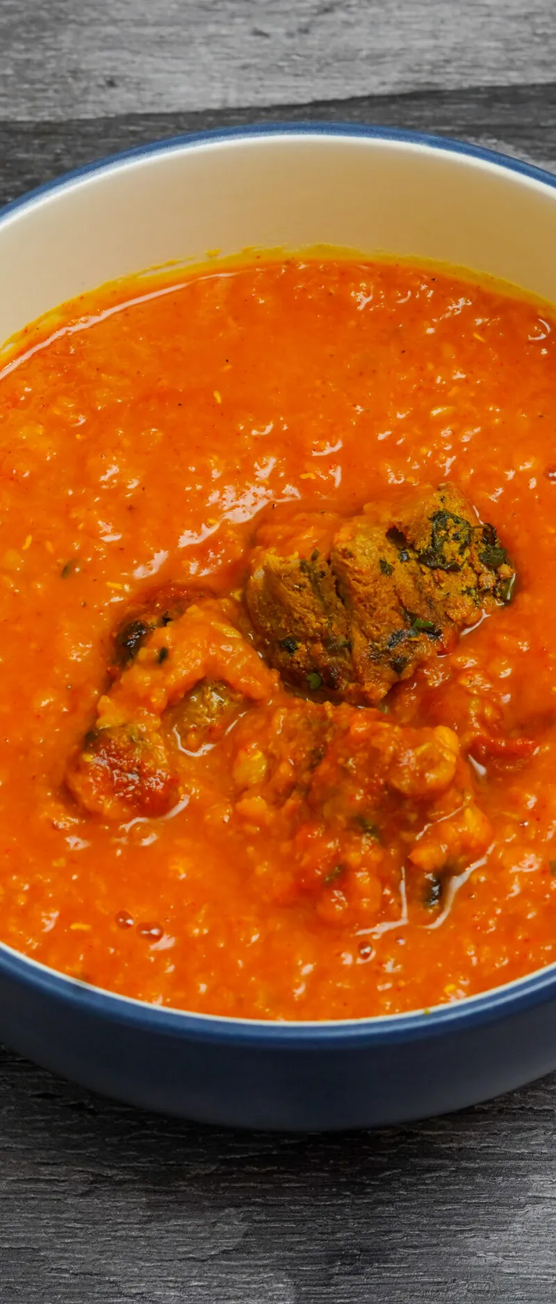 Bright orange lentil curry (dal) containing several kofta balls, served in a white bowl with a blue rim, on a dark wooden surface.