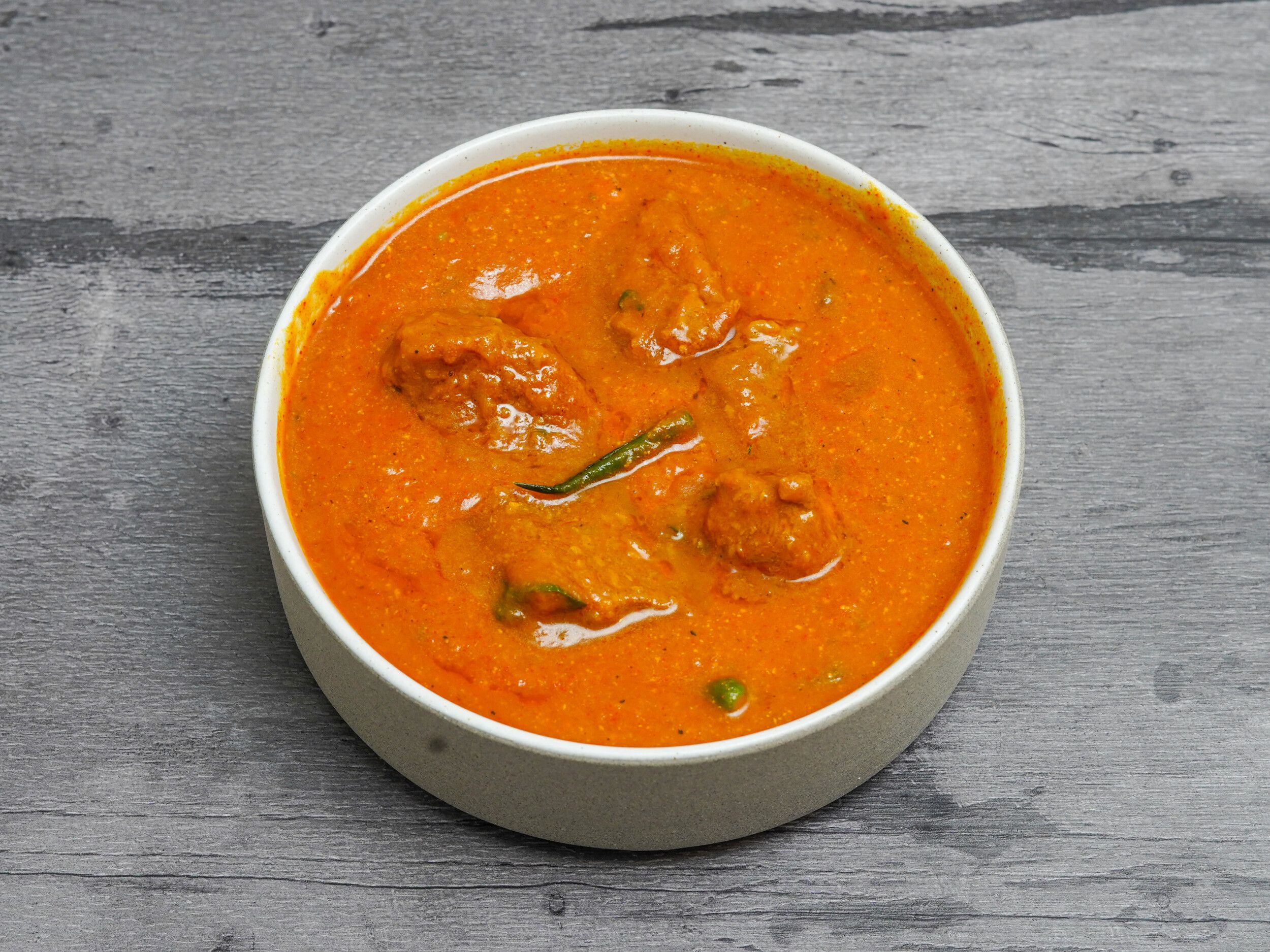 A white bowl filled with orange chicken tikka masala, garnished with a green chili.