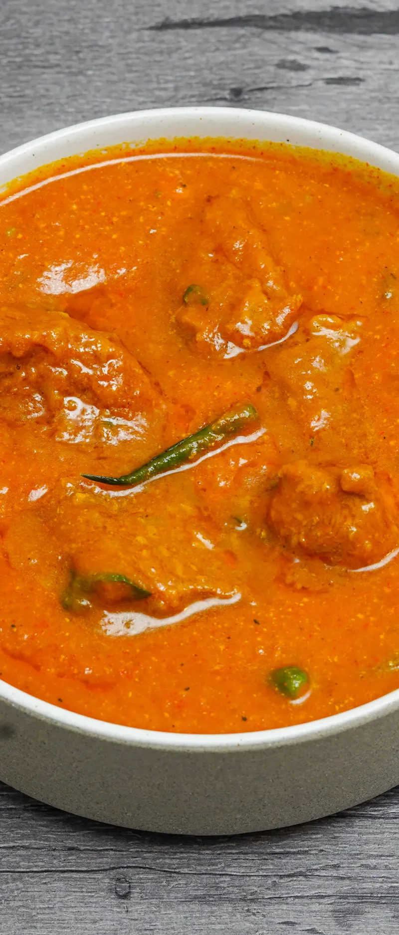 A white bowl filled with orange chicken tikka masala, garnished with a green chili.
