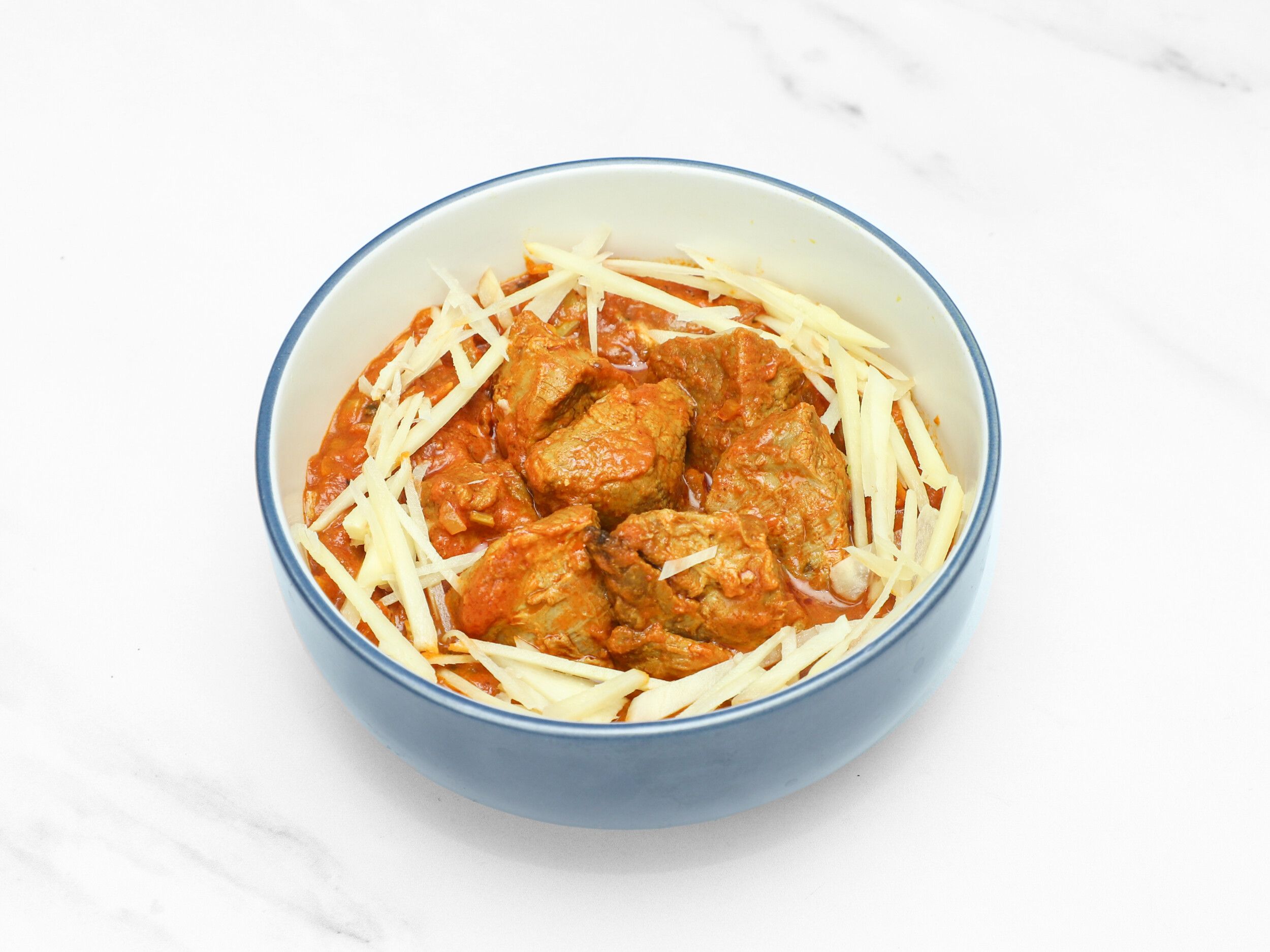 Lamb pieces in a thick red curry sauce, garnished with thin white ginger strips, served in a blue and white bowl