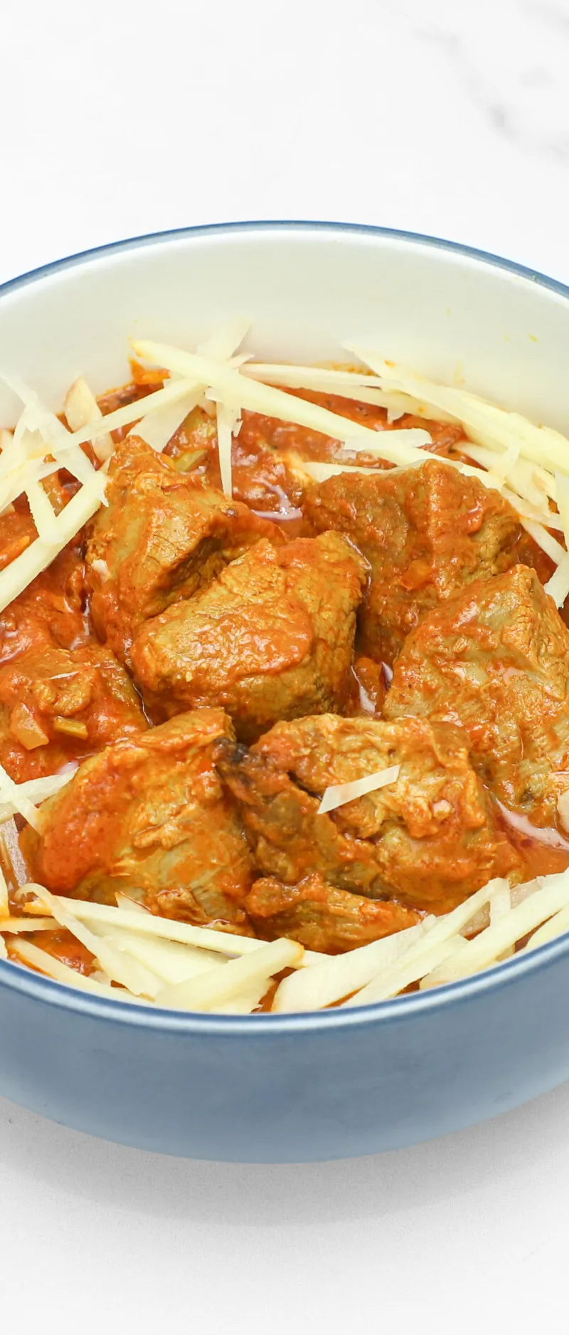 Lamb pieces in a thick red curry sauce, garnished with thin white ginger strips, served in a blue and white bowl