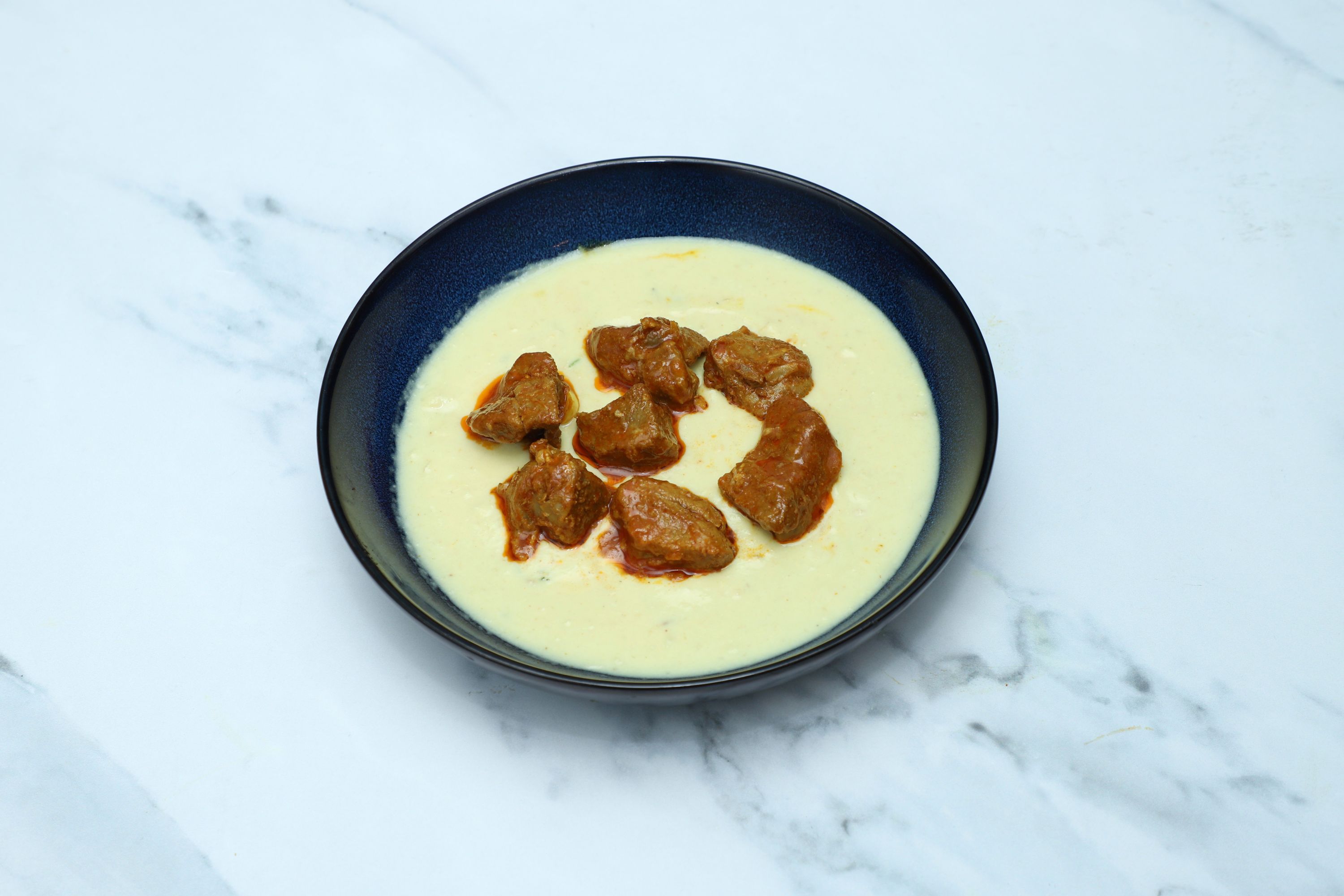 A round blue ceramic bowl filled with a creamy yellow curry and several chunks of meat.