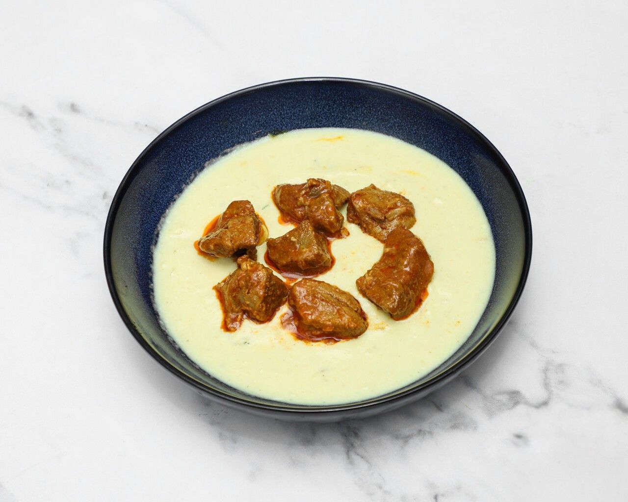 Top-down view of tender lamb bhuna pieces in a creamy, pale yellow sauce served in a blue bowl.