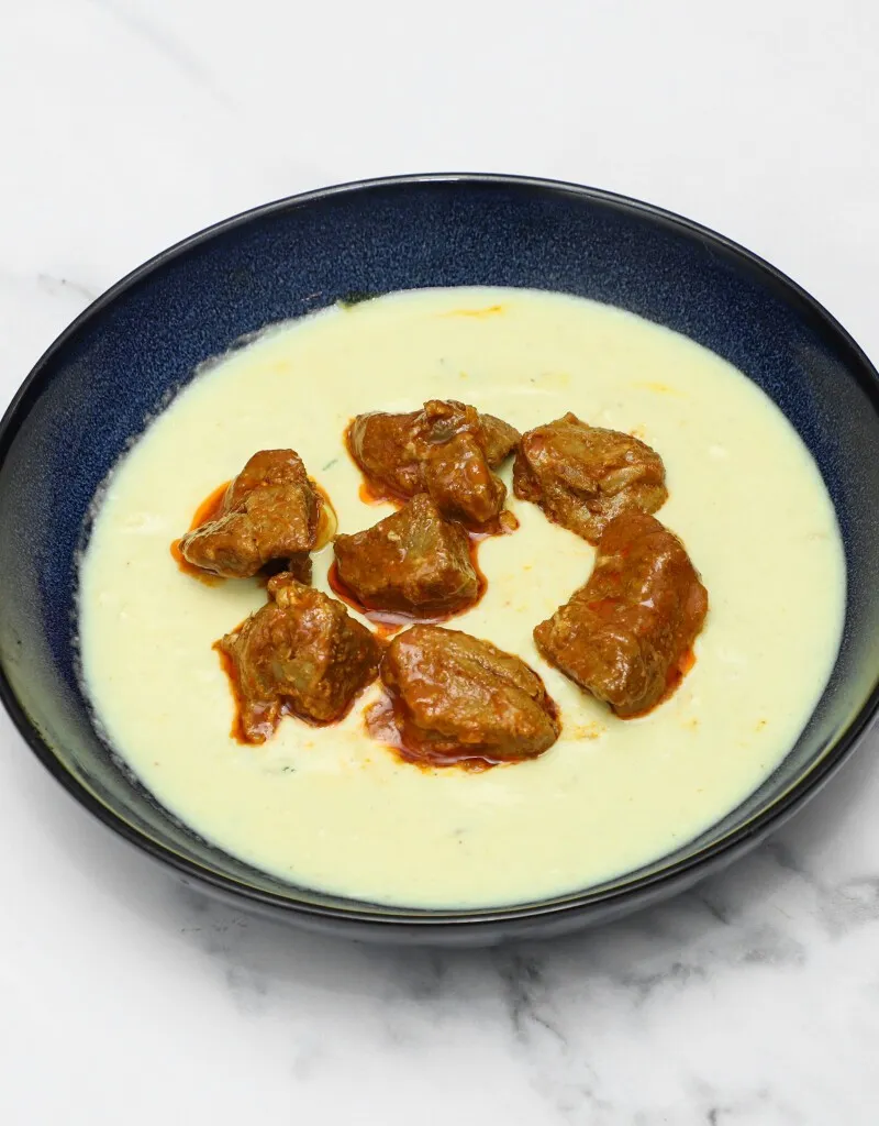 Top-down view of tender lamb bhuna pieces in a creamy, pale yellow sauce served in a blue bowl.