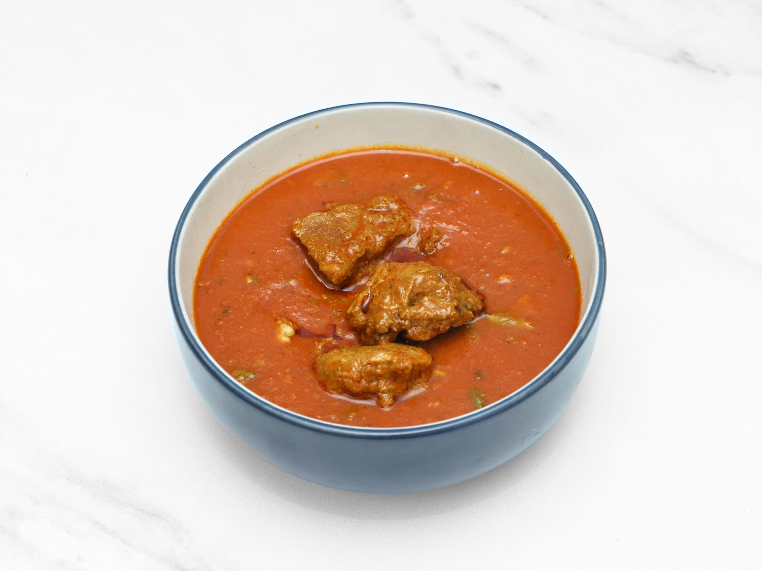 A blue and white bowl filled with a vibrant red curry and chunky pieces of meat on a white marble background.