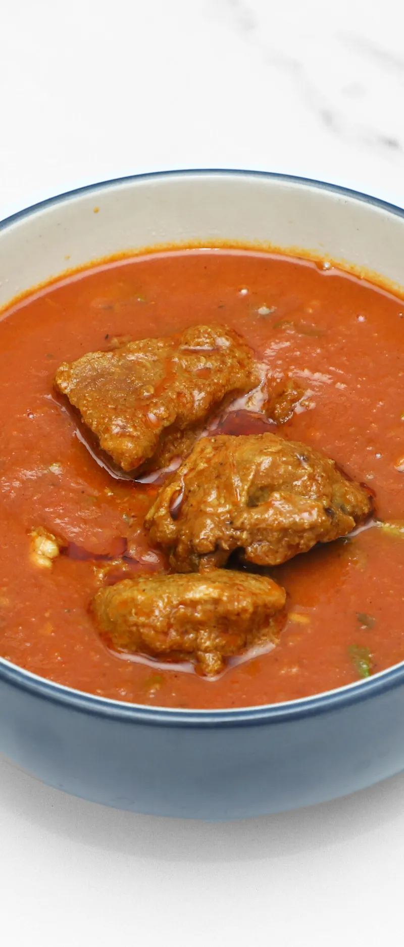 A blue and white bowl filled with a vibrant red curry and chunky pieces of meat on a white marble background.