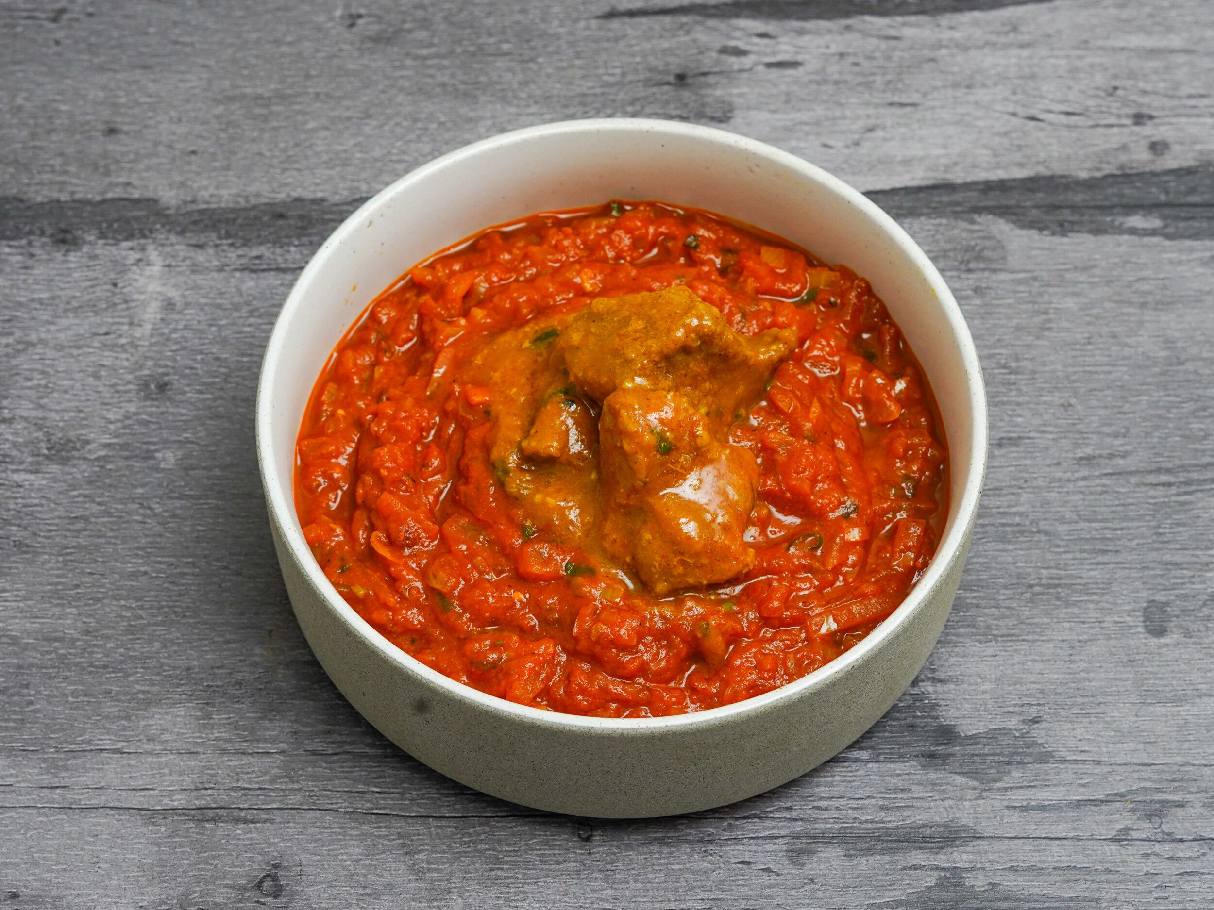 Chicken curry featuring pieces of chicken in a vibrant red tomato-based sauce, served in a white bowl.