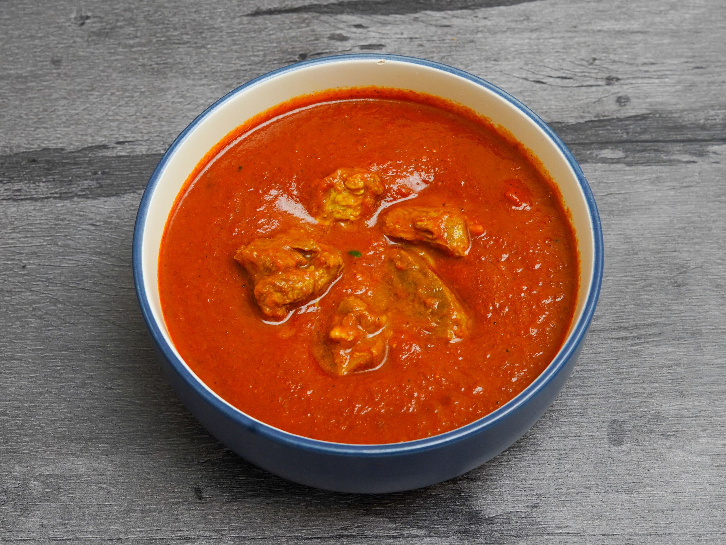 Chicken curry with chunks of meat in a vibrant red sauce, served in a blue and white ceramic bowl.