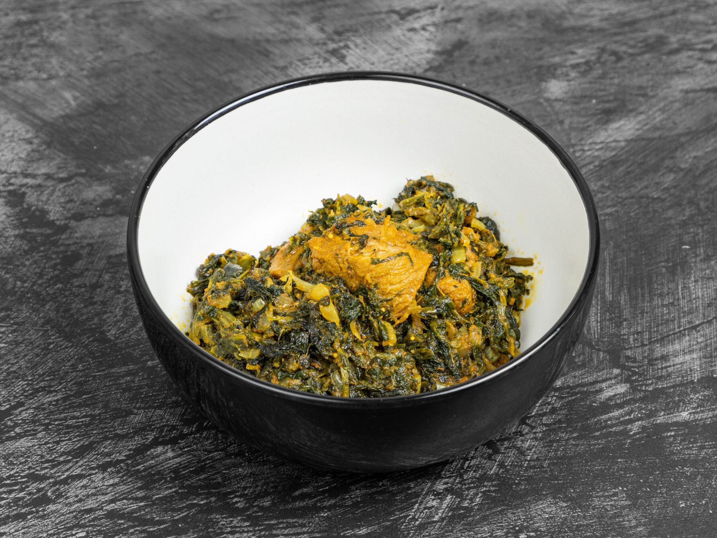 Sag chicken curry in a black and white bowl, on a dark textured surface