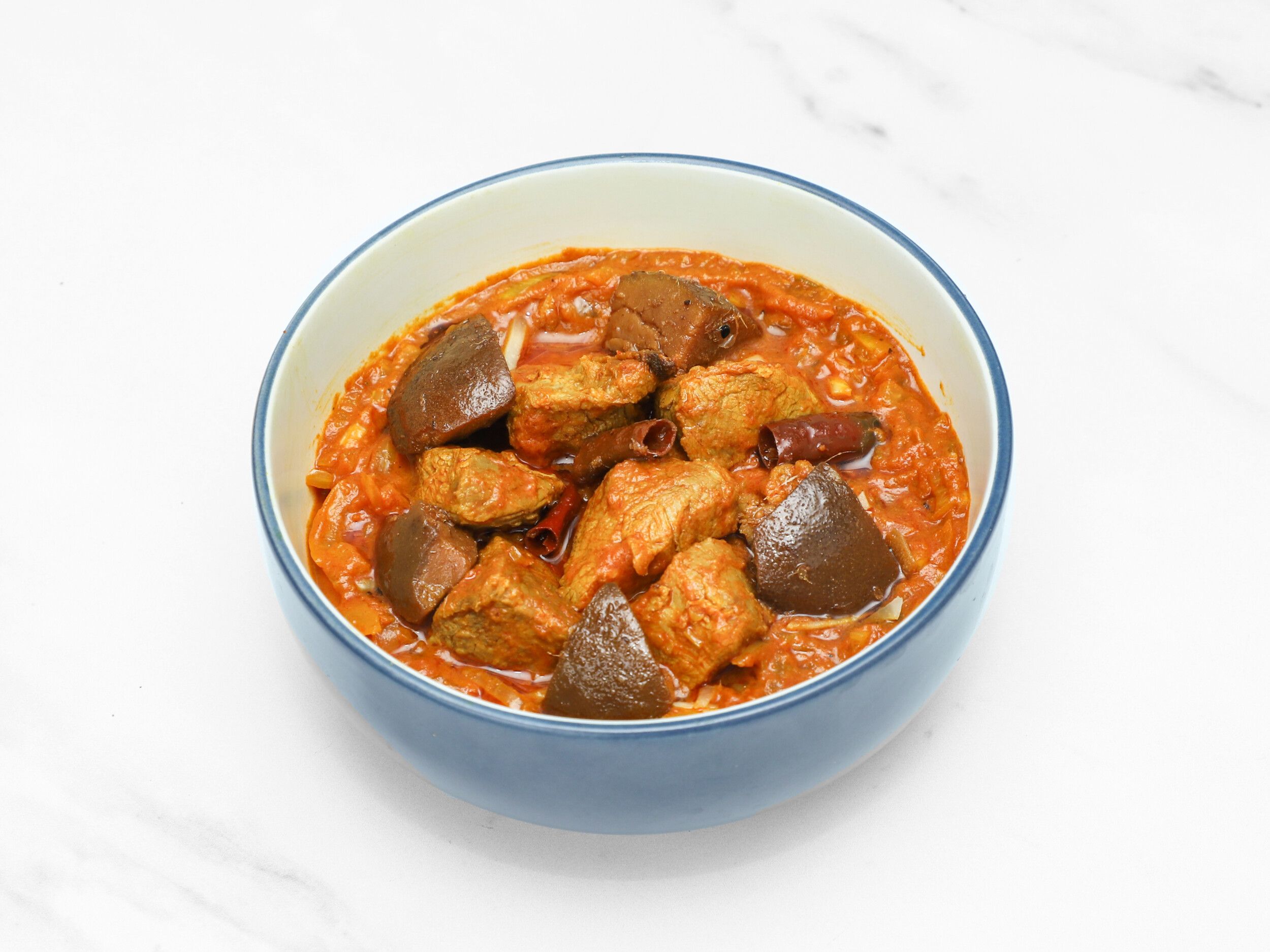 Meat and aubergine curry in a blue and white ceramic bowl on a white surface.