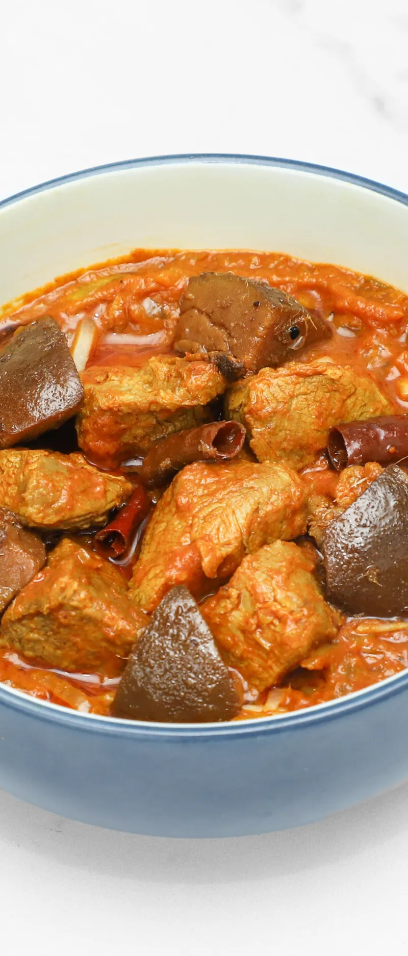 Meat and aubergine curry in a blue and white ceramic bowl on a white surface.