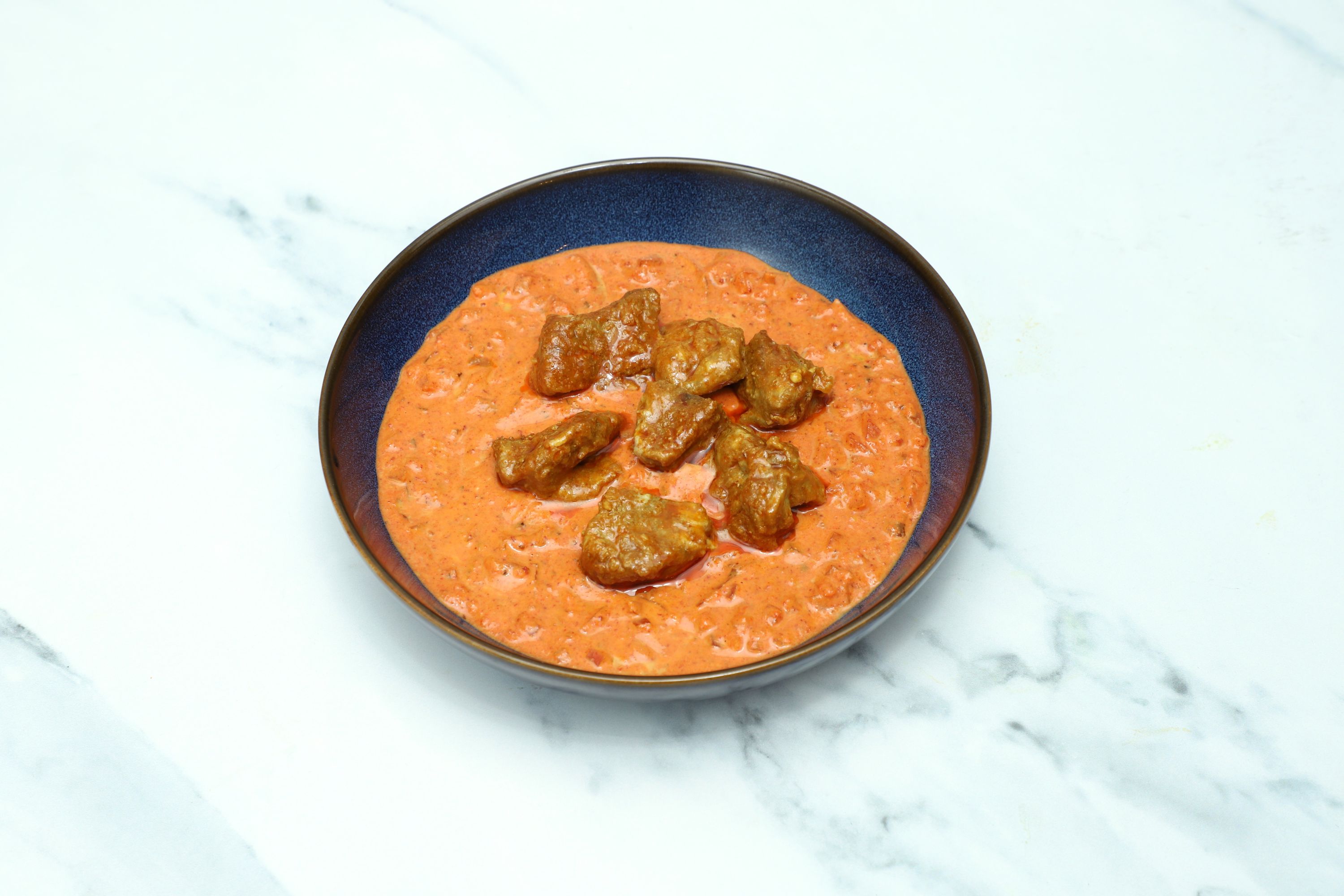 A dark blue bowl of lamb bhuna, featuring tender meat pieces in a rich, orange-red creamy sauce, on a white marble surface.
