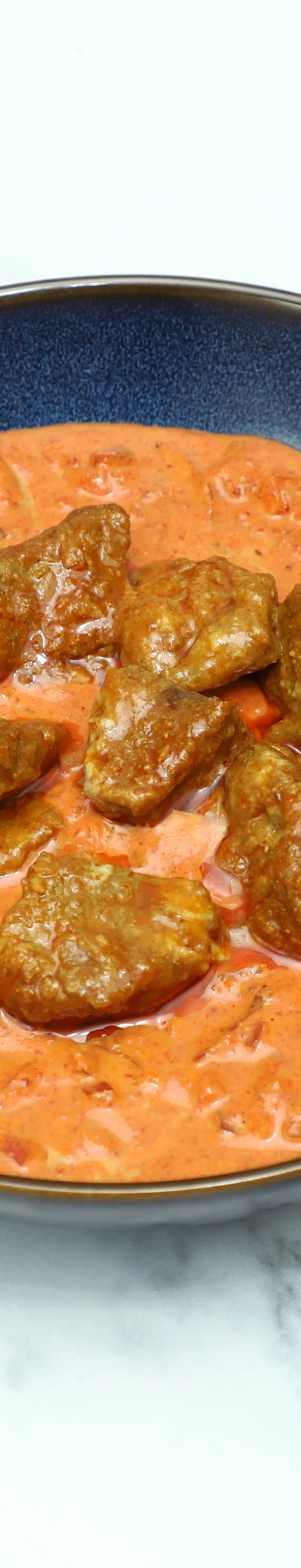 A dark blue bowl of lamb bhuna, featuring tender meat pieces in a rich, orange-red creamy sauce, on a white marble surface.